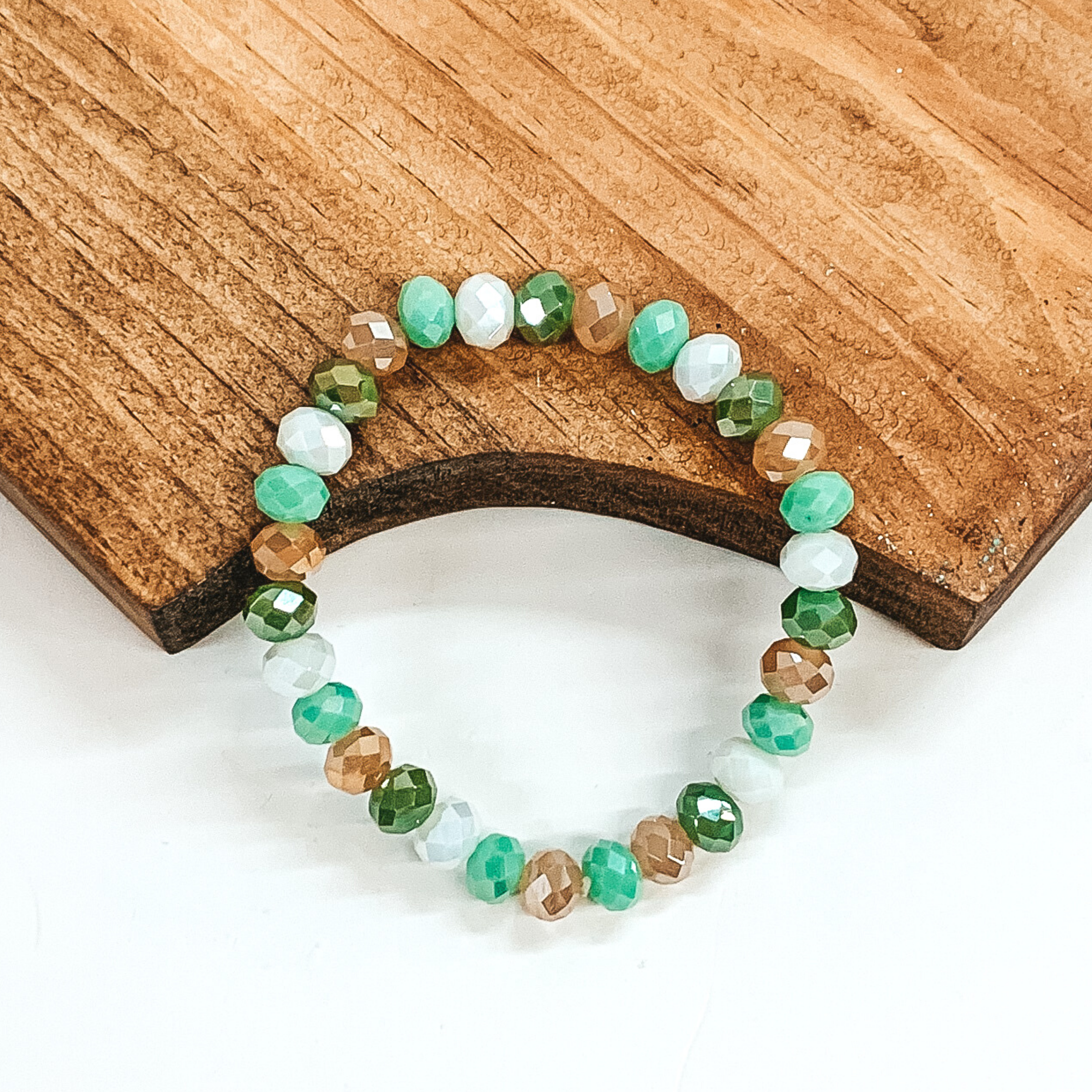 Crystal beaded bracelet in ivory, light green, green, and light brown. This bracelet is pictured on a partially laying on a brown block on a white background.