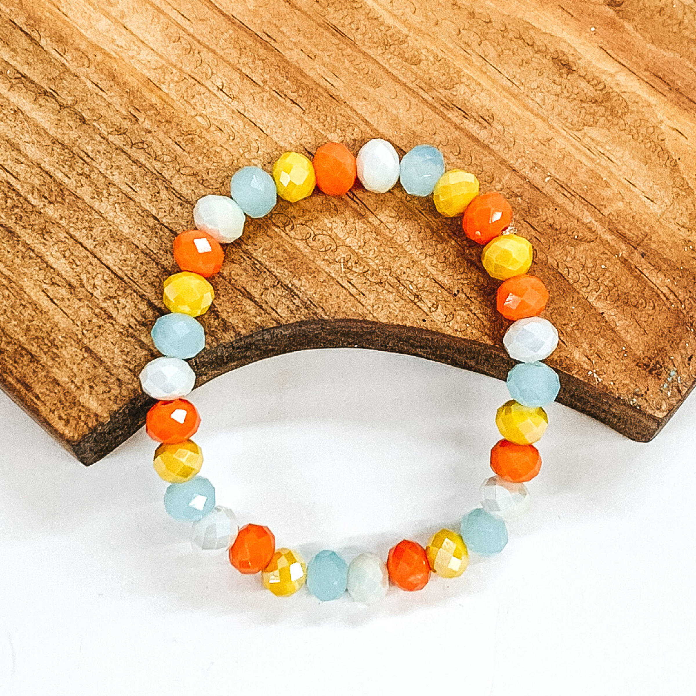 Crystal beaded bracelet in white, yellow, orange, and pale blue. This bracelet is pictured on a partially laying on a brown block on a white background.