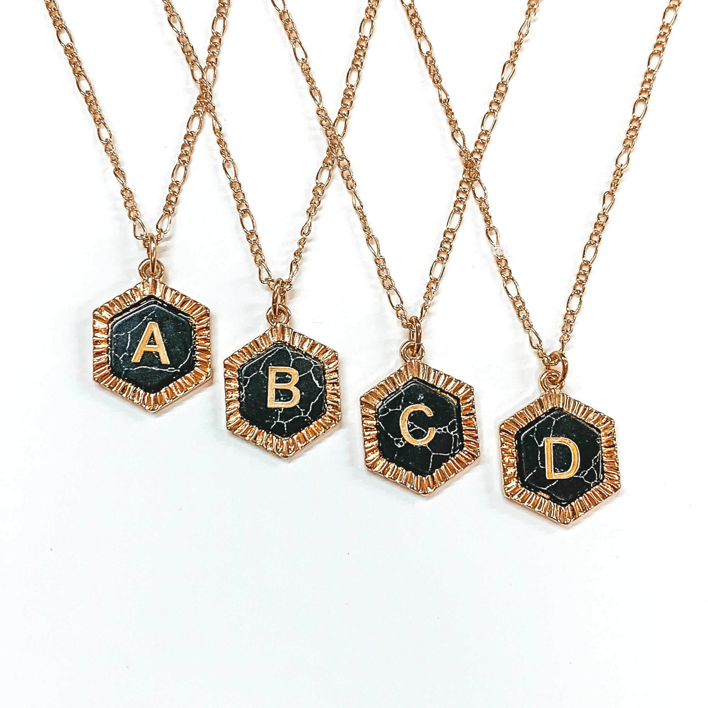 Four gold, figaro chained necklaces with hexagon pendants. Each pendant is outlined with gold and has a black stone center with a gold initial. This picture includes the initials "A, B, C, and D." These necklaces are pictured on a white background.