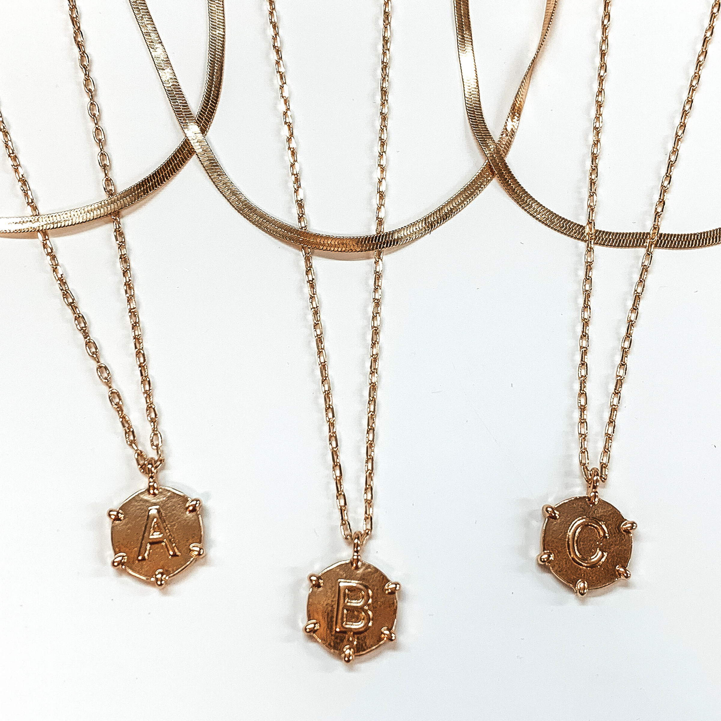 Three gold, snake chain and paperclip chain necklaces with circle pendants. Each pendant has gold beads outlining the pendant with a centered gold initial. This picture includes the initials "A, B, and C." These necklaces are pictured on a white background.