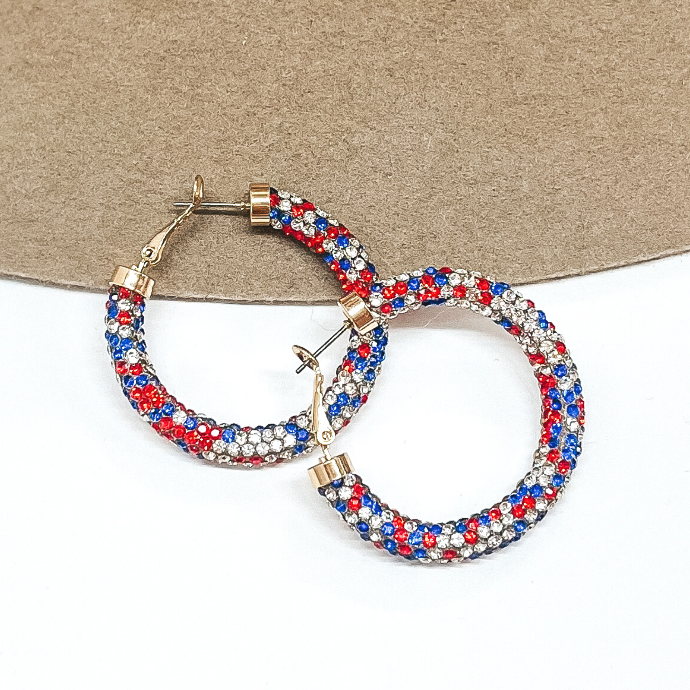 Hoop earrings with a rhinstone mesh covering that includes red, white, and blue colors. These hoops have gold ends. These earrings are pictured on a brown and white background.