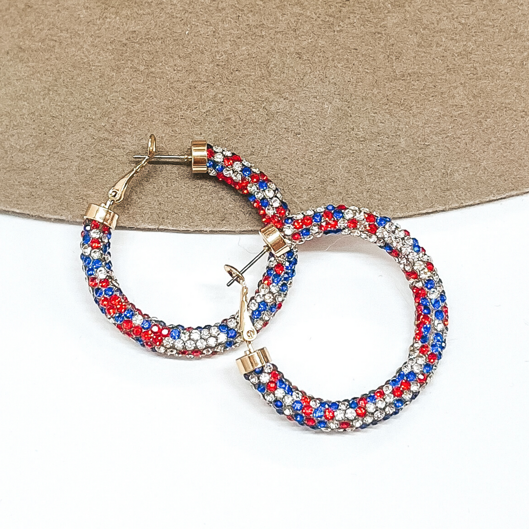 Hoop earrings with a rhinstone mesh covering that includes red, white, and blue colors. These hoops have gold ends. These earrings are pictured on a brown and white background.