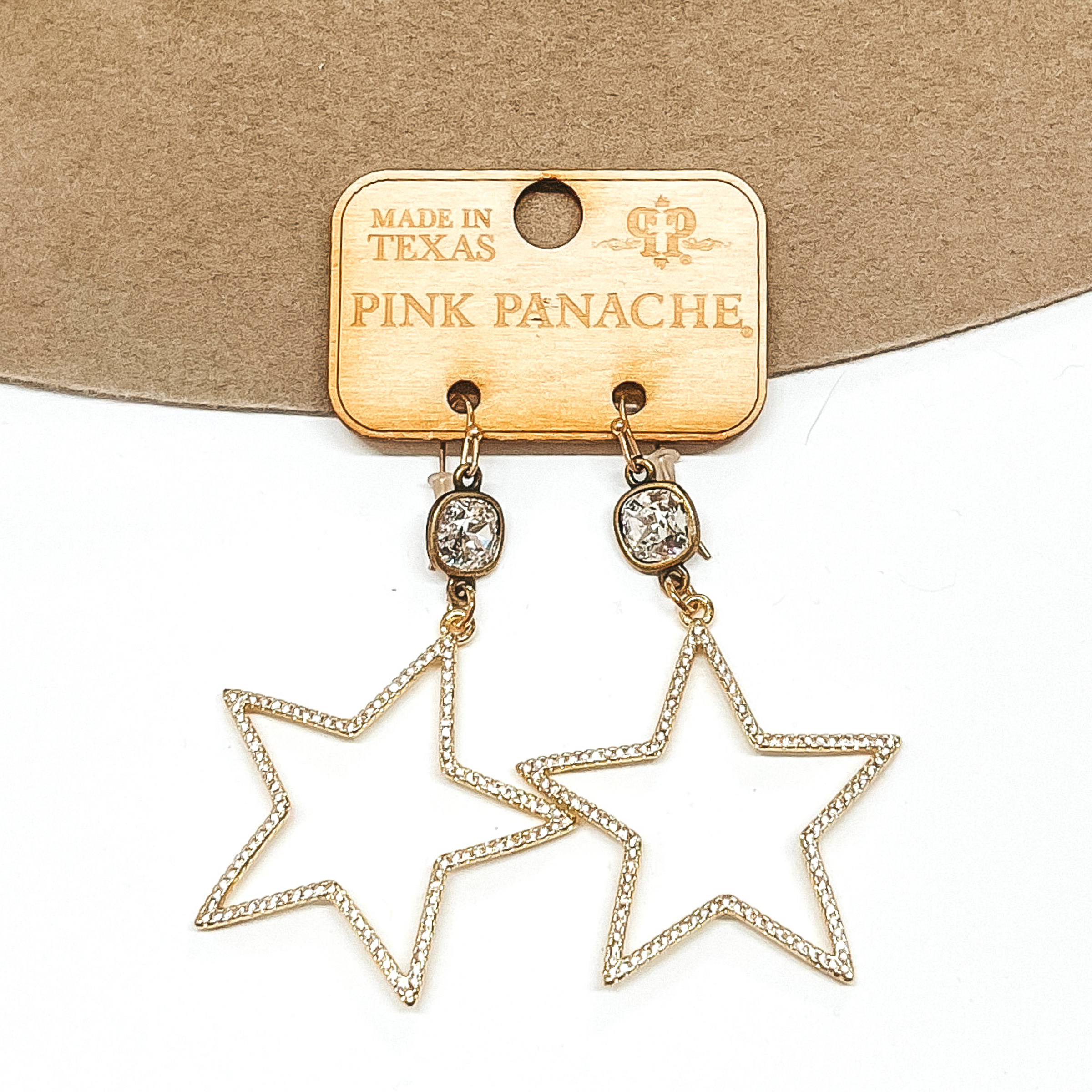 Clear cushion cut crystals that are in a bronze settings and are connected to bronze fish hook earrings. Hanging from the crystals are gold open star pendants. These earrings are pictured on a tan and white background. 