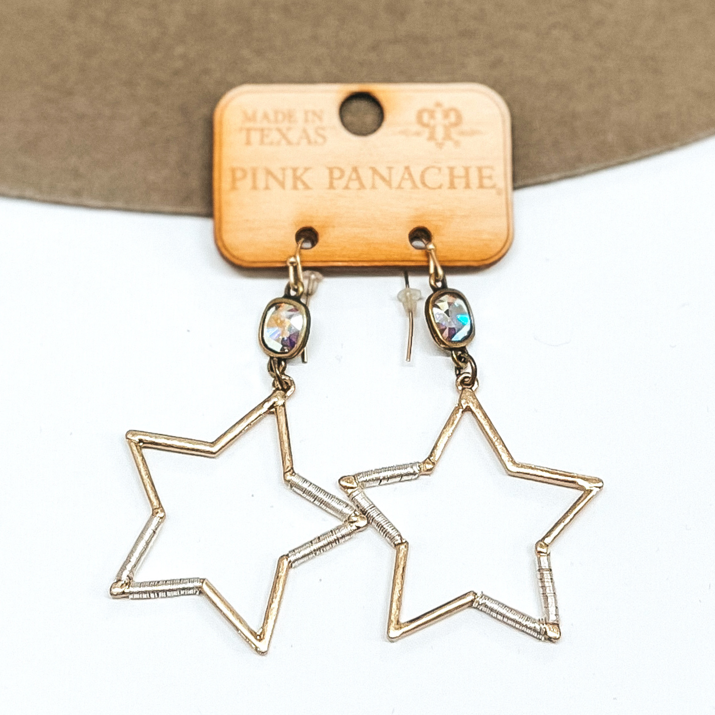 AB cushion cut crystals that are in a bronze settings and are connected to bronze fish hook earrings. Hanging from the crystals are gold open star pendant with silver wire wrapping around a few sides. These earrings are pictured on a tan and white background.