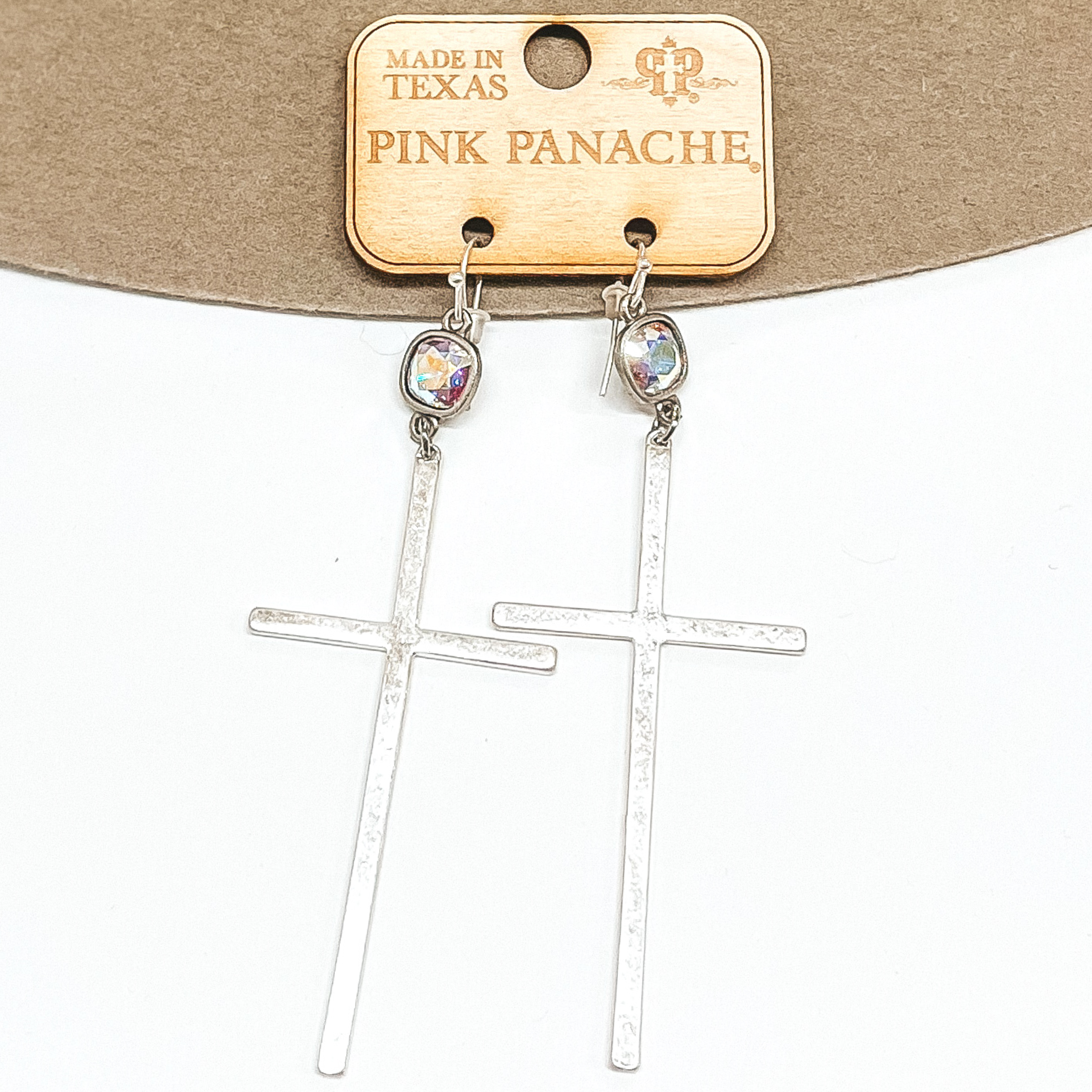 AB cushion cut crystals that are in a silver setting and are connected to silver fish hook earrings. Hanging from the crystals are smooth, silver cross pendants. These earrings are pictured on a tan and white background.