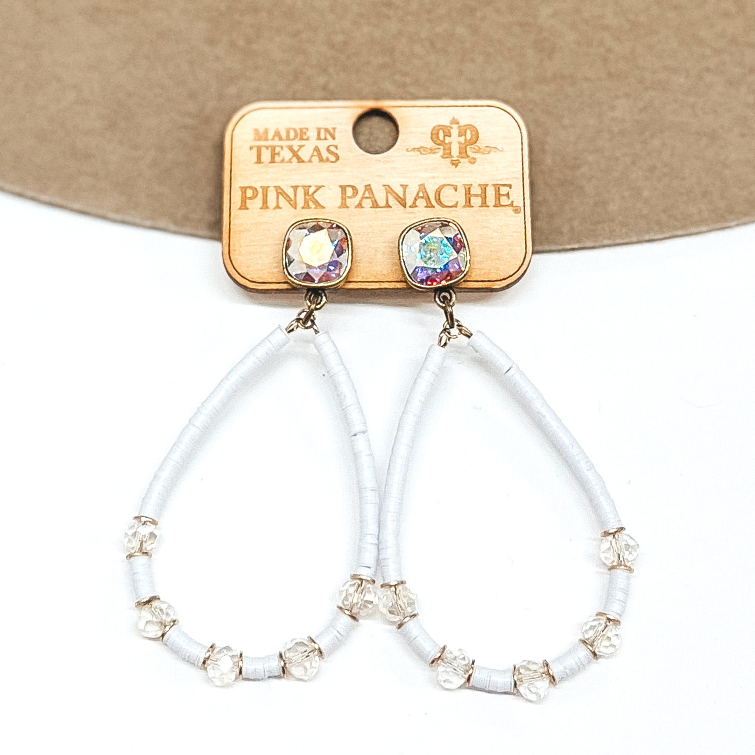 AB cushion cut crystal earrings in a bronze setting. These earrings include a teardrop pendant that has white, rubber disk beads and clear crystal spacers. These earrings are pictured on a tan and white background. 