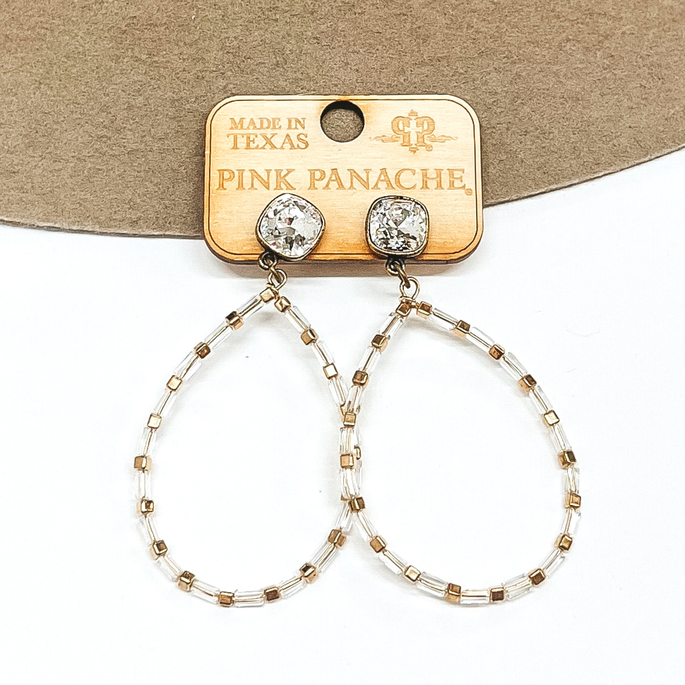 Clear cushion cut crystal earrings in a bronze setting. These earrings include a teardrop pendant that has clear beads with gold bead spacers. These earrings are pictured on a tan and white background.