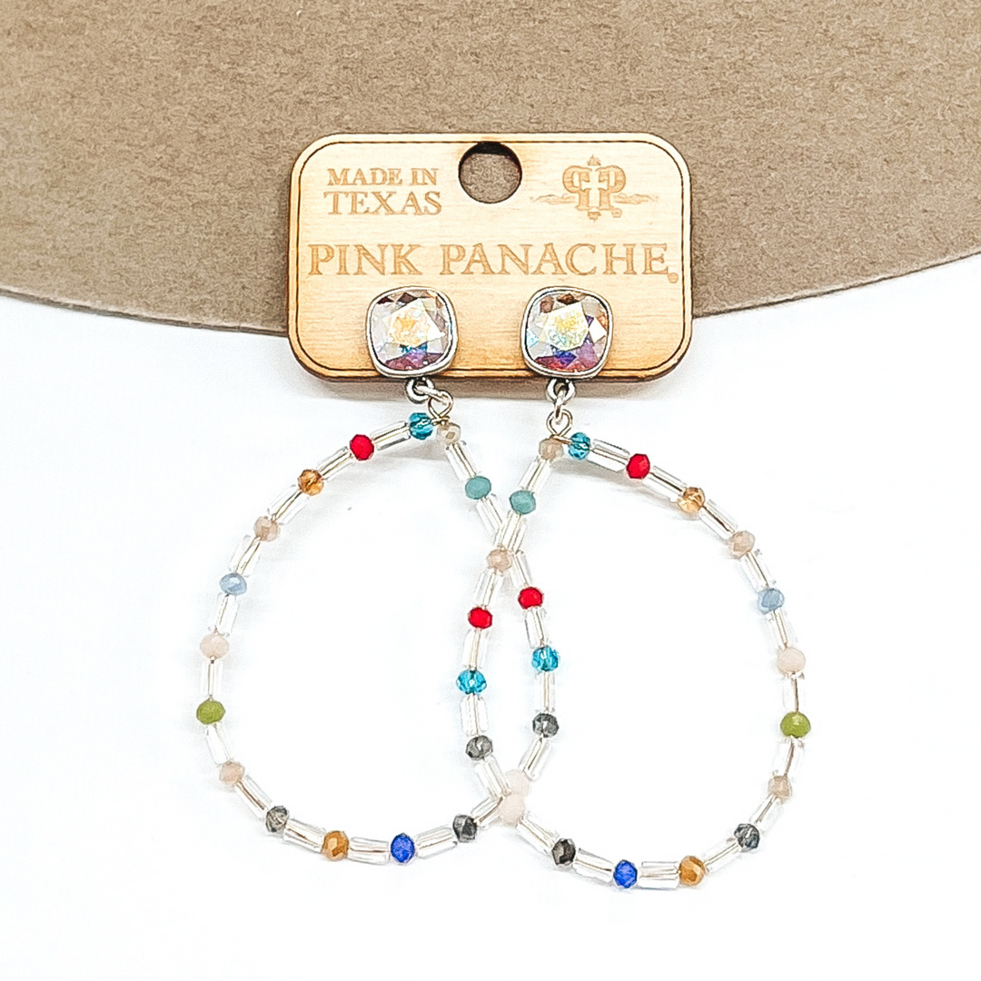 AB cushion cut crystal earrings in a silver setting. These earrings include a teardrop pendant that has clear beads with multicolored bead spacers. These earrings are pictured on a tan and white background.