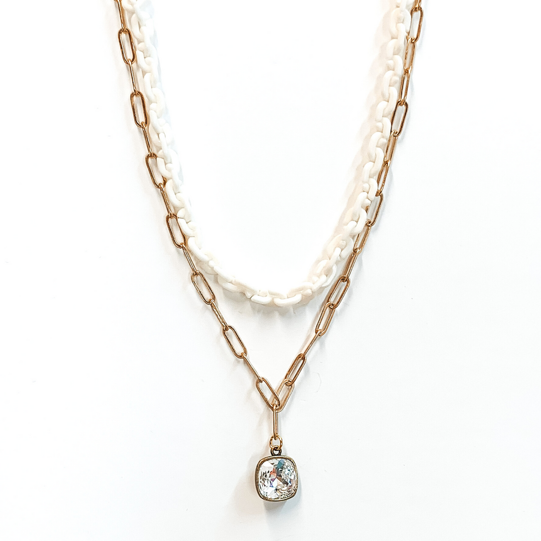 Two strand chain necklace. The shorter chain is a thick white chain. The longer chain is a simple gold paperclip chain with a hanging clear cushion cut crystal pendant with a bronze backing. This necklace is pictured on a white background.