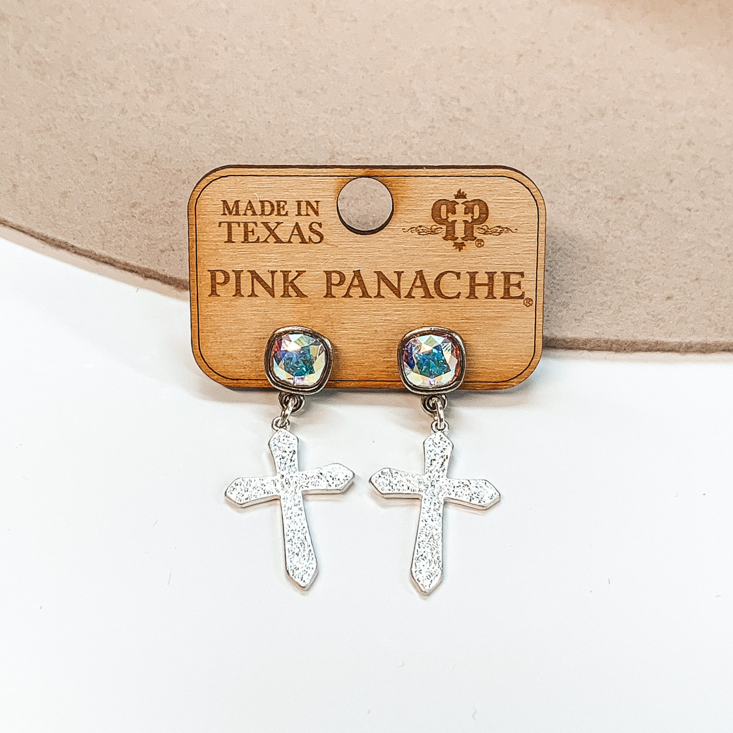 Ab cushion cut crystal stud earrings with a hanging silver, textured cross pendant in silver. These earrings are pictured on a white and tan background. 