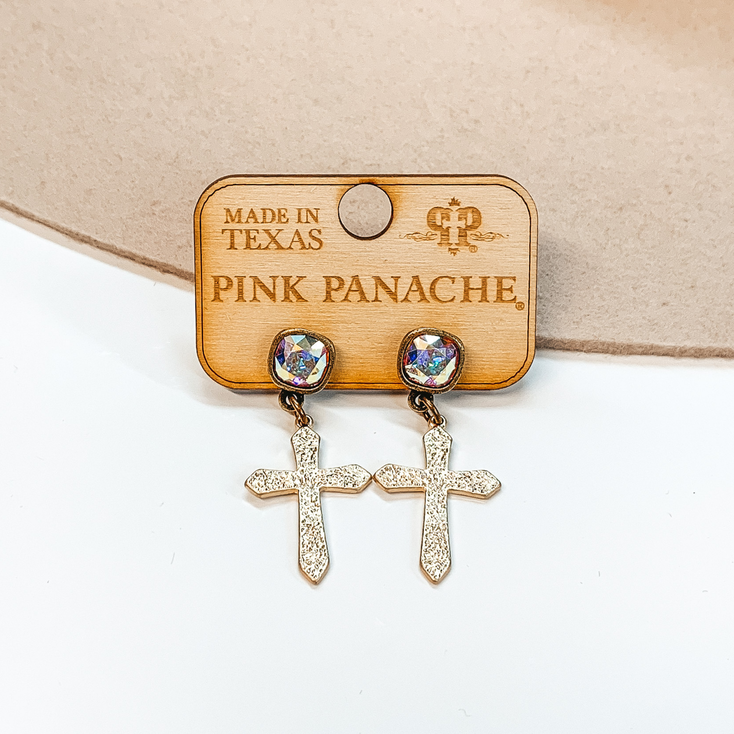Ab cushion cut crystal stud earrings with a hanging gold, textured cross pendant. These earrings are pictured on a white and tan background. 