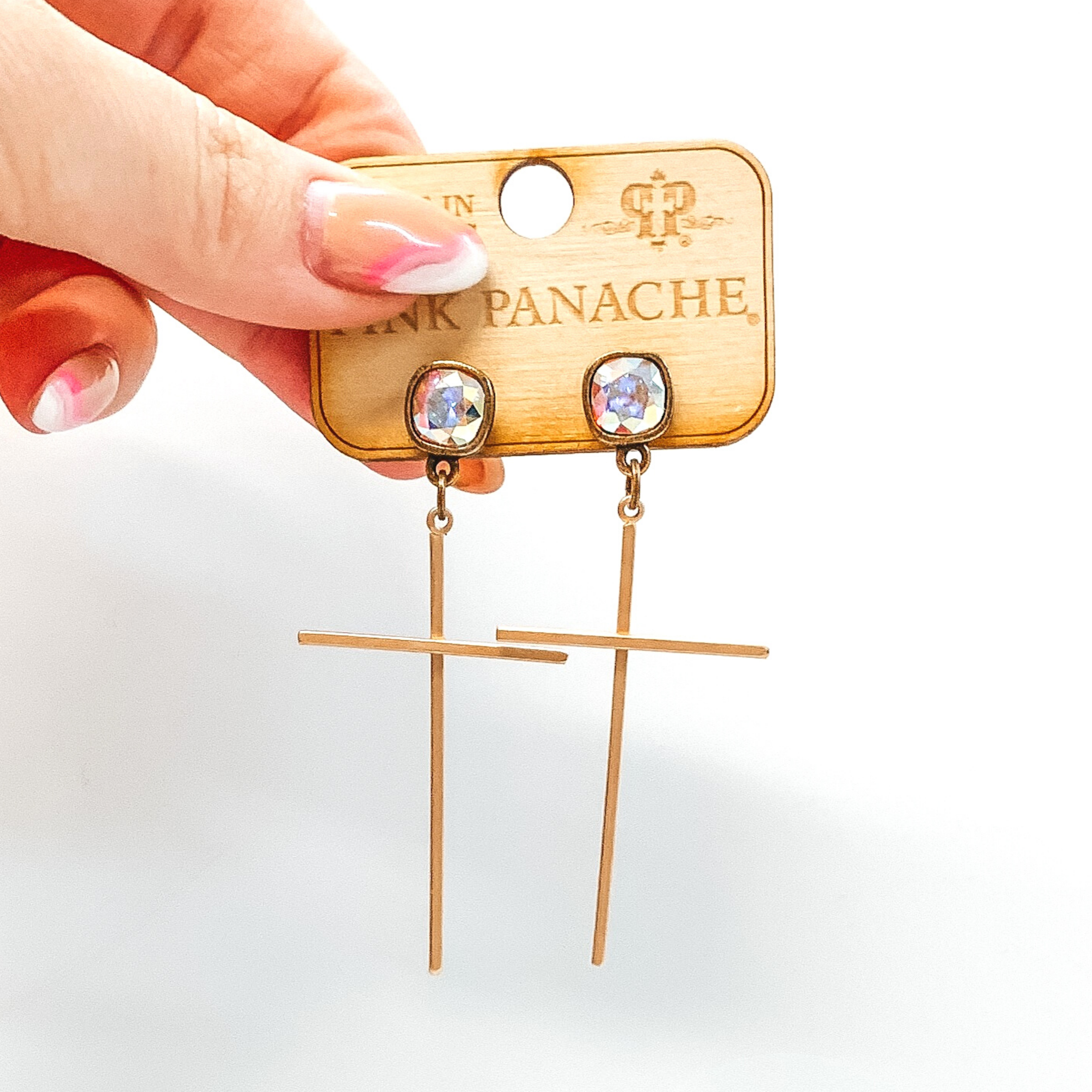 Ab cushion cut crystal stud earrings with a hanging gold, thin cross pendant. These earrings are pictured on a white background and is held up by a hand. 