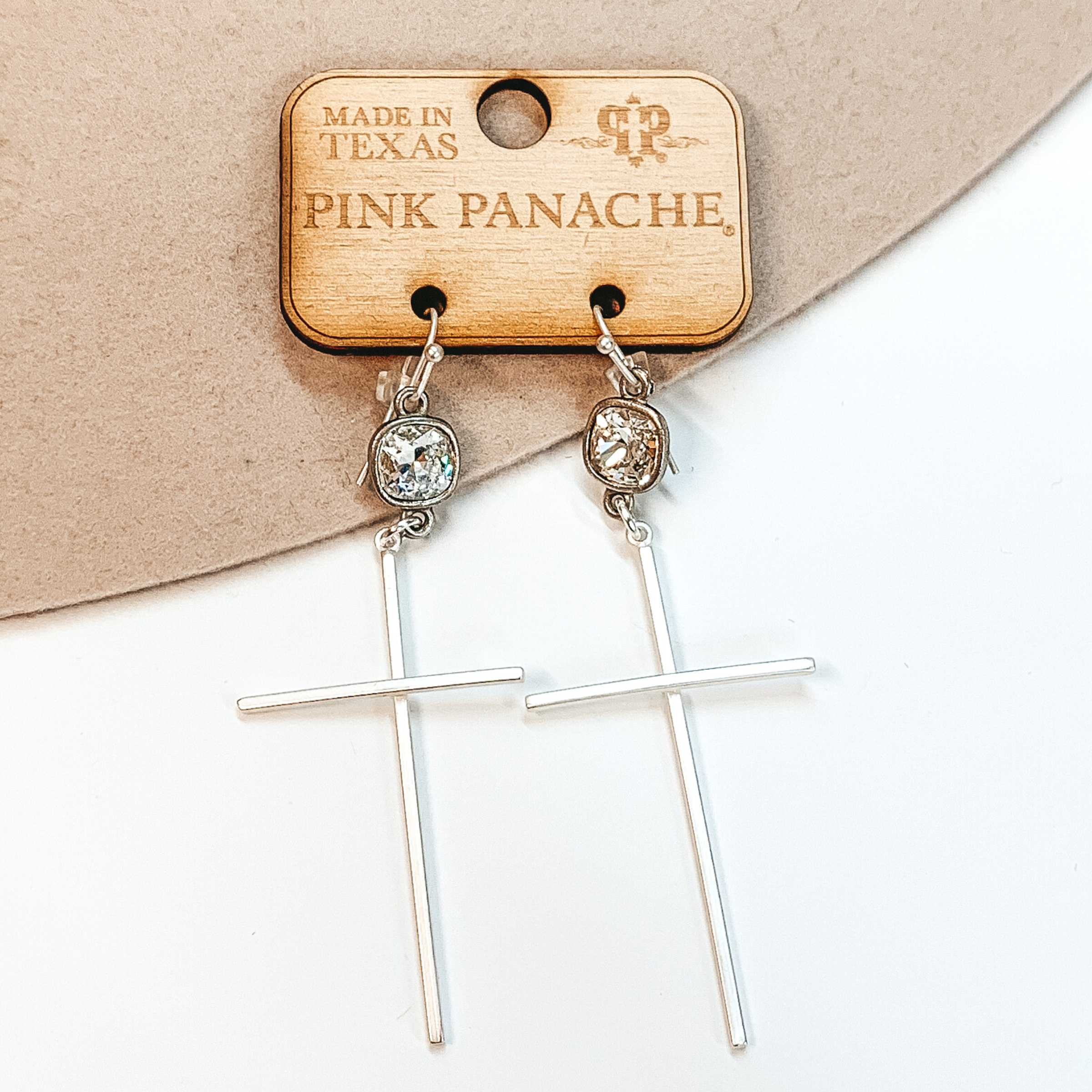 Clear cushion cut crystals on a silver fish hook earring with a hanging silver, thin cross pendant. These earrings are pictured on a white background and is held up by a hand. 