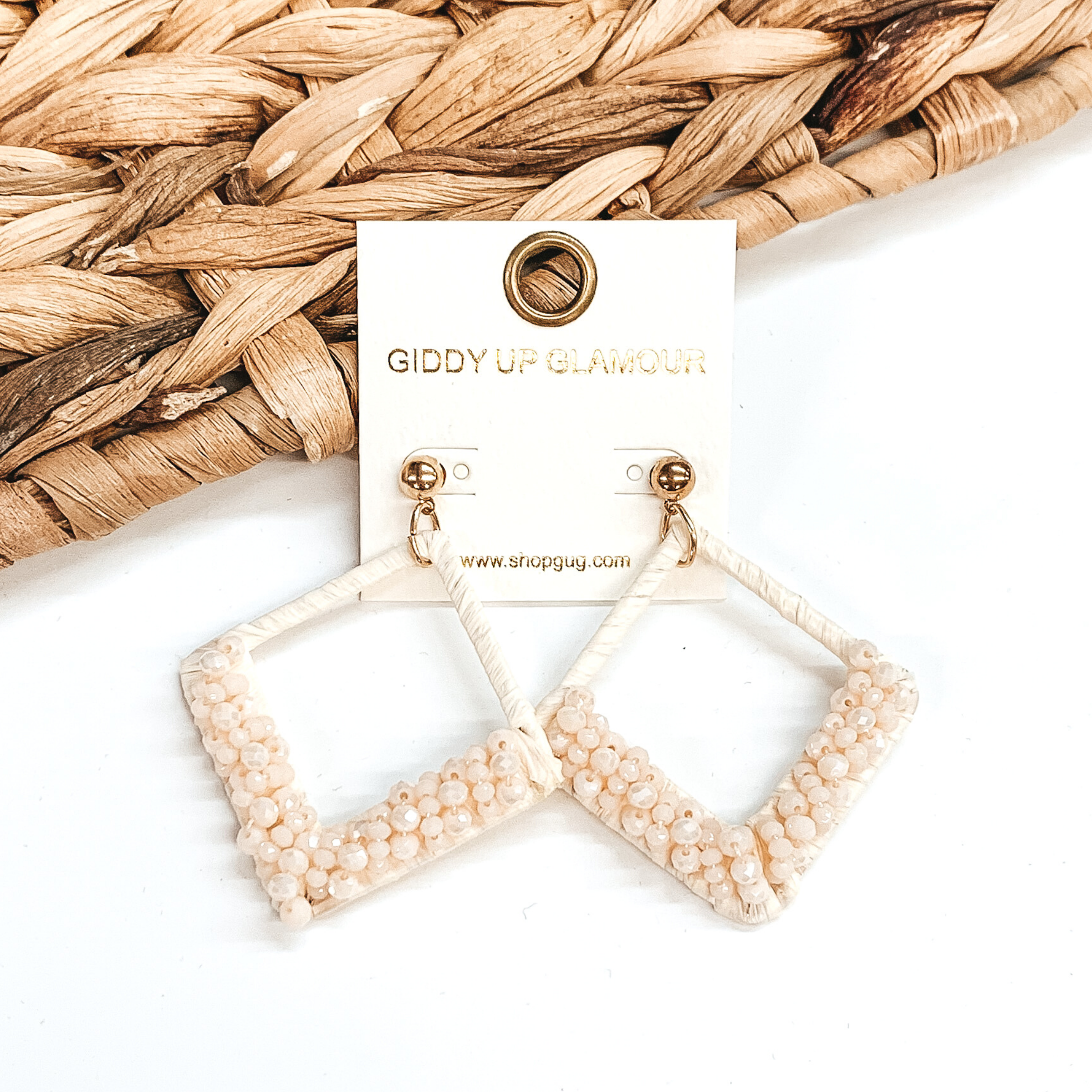  Open diamond shaped dangle earrings wrapped in raffia that includes a layer of beads on the bottom half of the earrings. These earrings are ivory in color. These earrings are pictured on on a white background with basket weave decor.