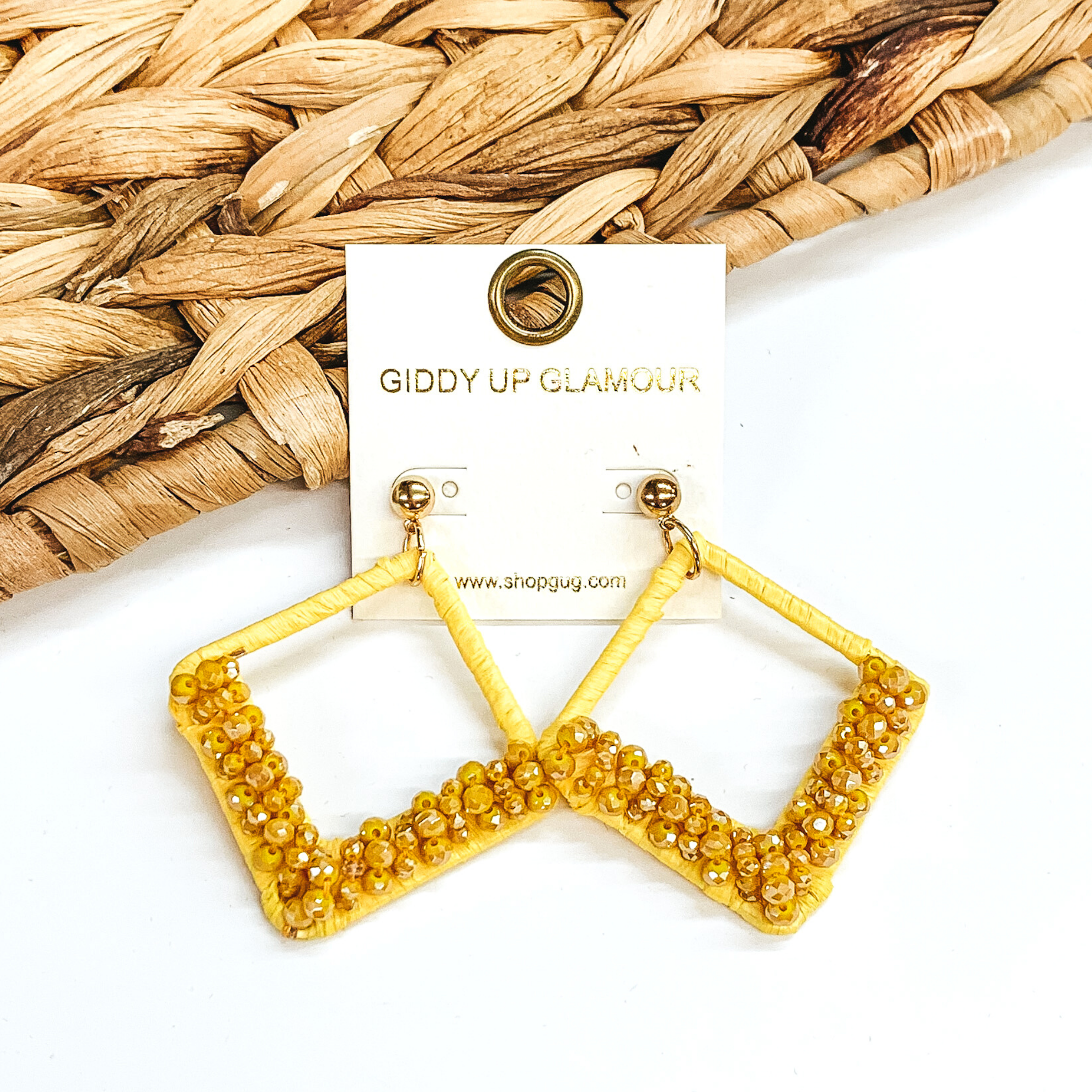 Open diamond shaped dangle earrings wrapped in raffia that includes a layer of beads on the bottom half of the earrings. These earrings are yellow in color. These earrings are pictured on on a white background with basket weave decor.