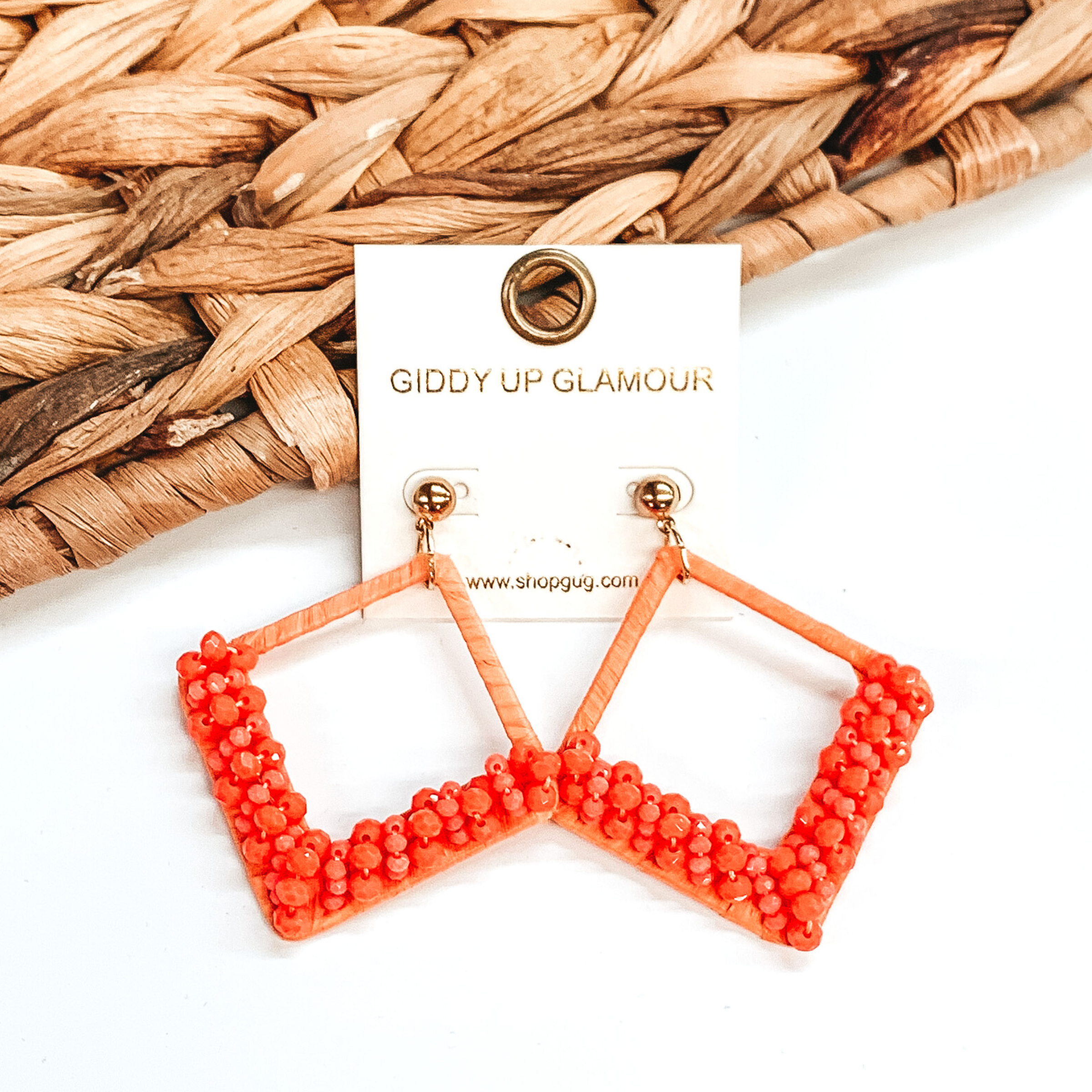 Open diamond shaped dangle earrings wrapped in raffia that includes a layer of beads on the bottom half of the earrings. These earrings are orange in color. These earrings are pictured on on a white background with basket weave decor.
