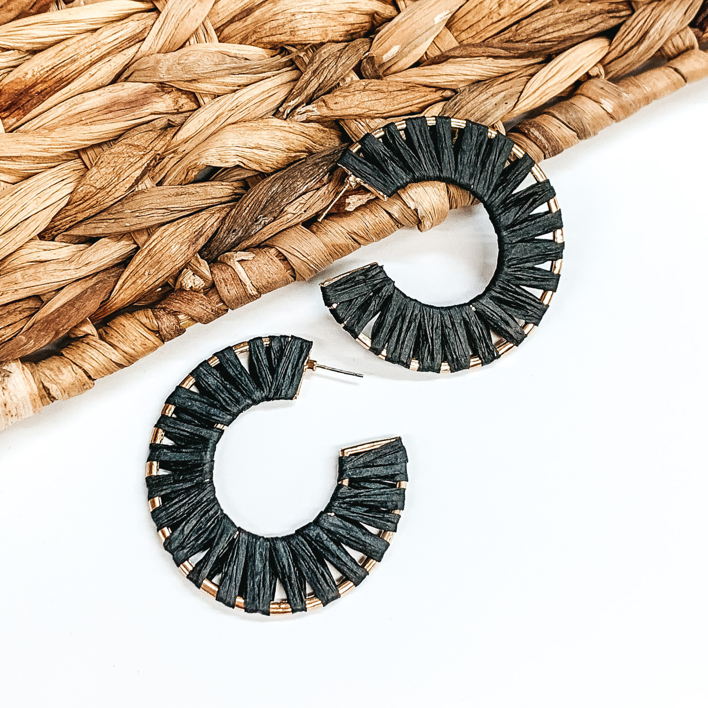 Gold wire circle hoop earrings that are wrapped in black raffia. These earrings are pictured on a white background with one of the hoops leaning on tan basket weave material.