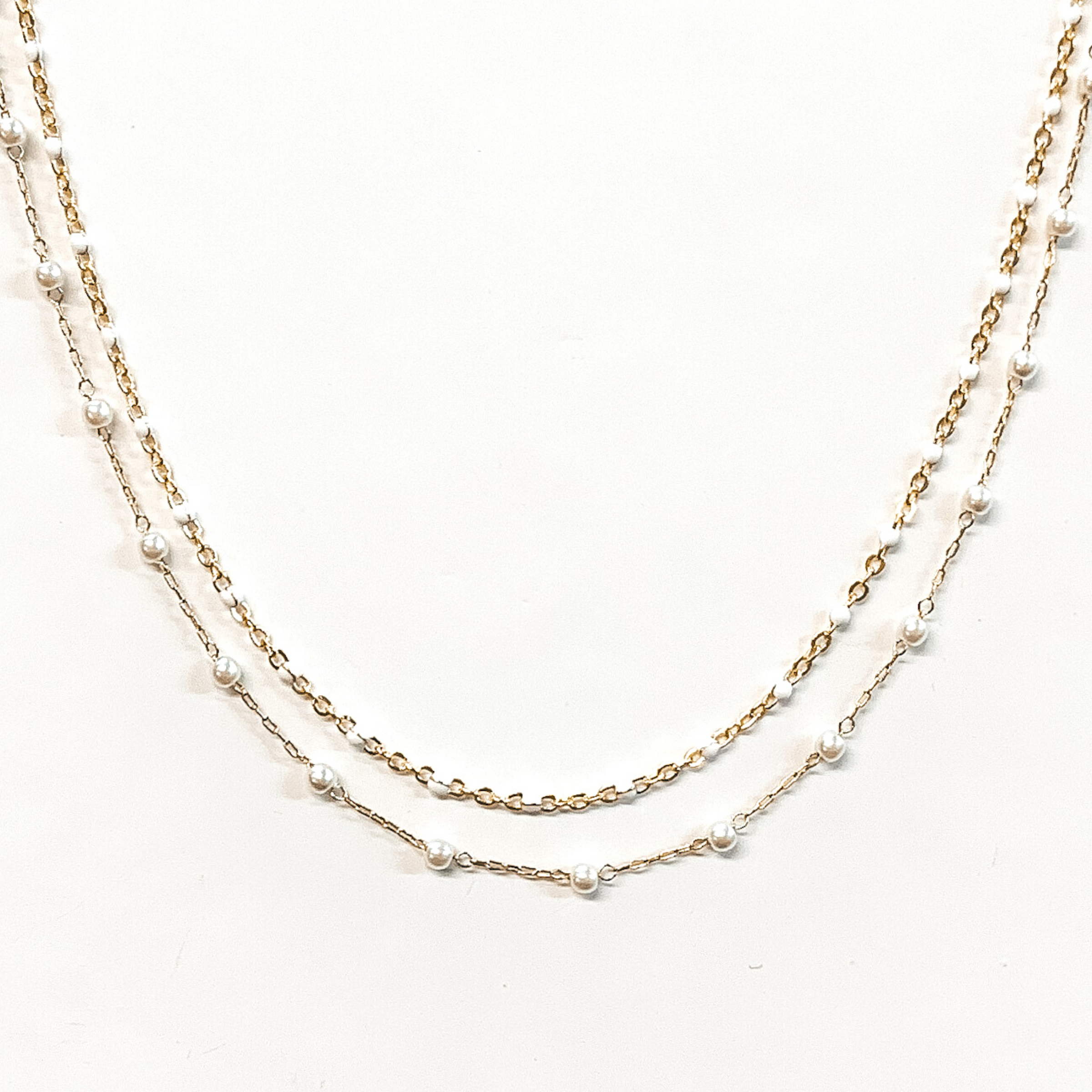 The first strand is a gold chain with small, white bead spacers. The second gold chain has small, white pearl bead spacers. This necklace is pictured on a white background.