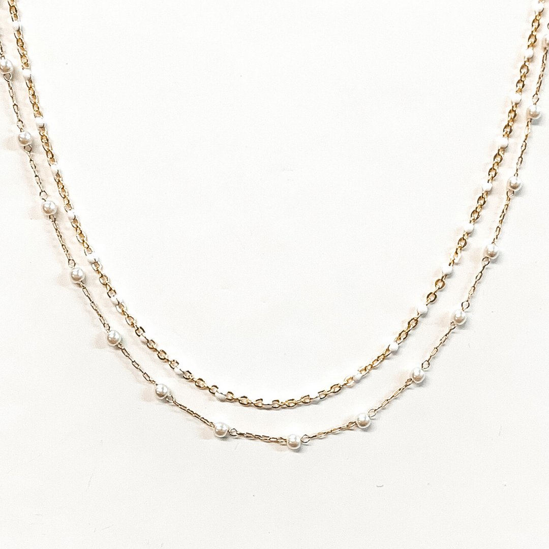 The first strand is a gold chain with small, white bead spacers. The second gold chain has small, white pearl bead spacers. This necklace is pictured on a white background.
