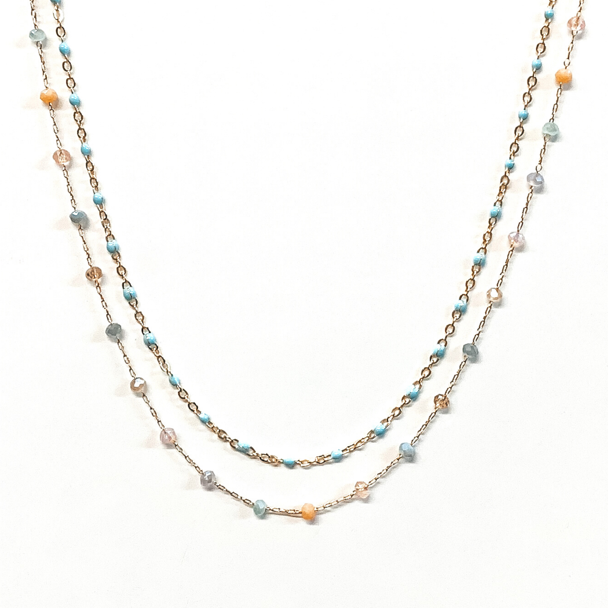 The first strand is a gold chain with small, baby blue bead spacers. The second gold chain has small, crystal bead spacers in a mix light blue, pink, and tan. This necklace is pictured on a white background.
