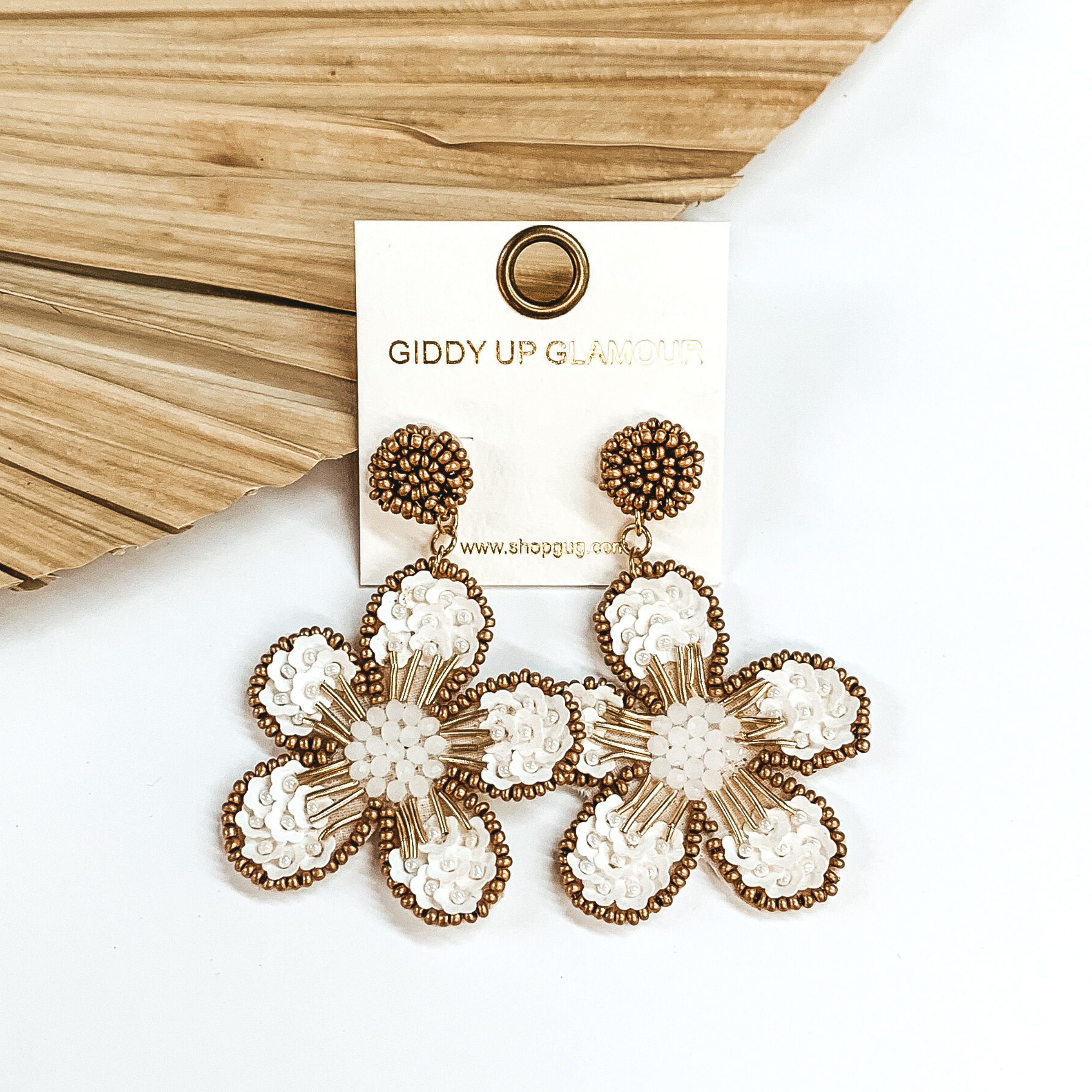Gold beaded circle post back earrings. There is a hanging beaded flower pendant from the gold stud earrings. The flower pendant is white with a gold outline and detailing. These earrings are pictured on a white background with a dried palm leaf in the background. 