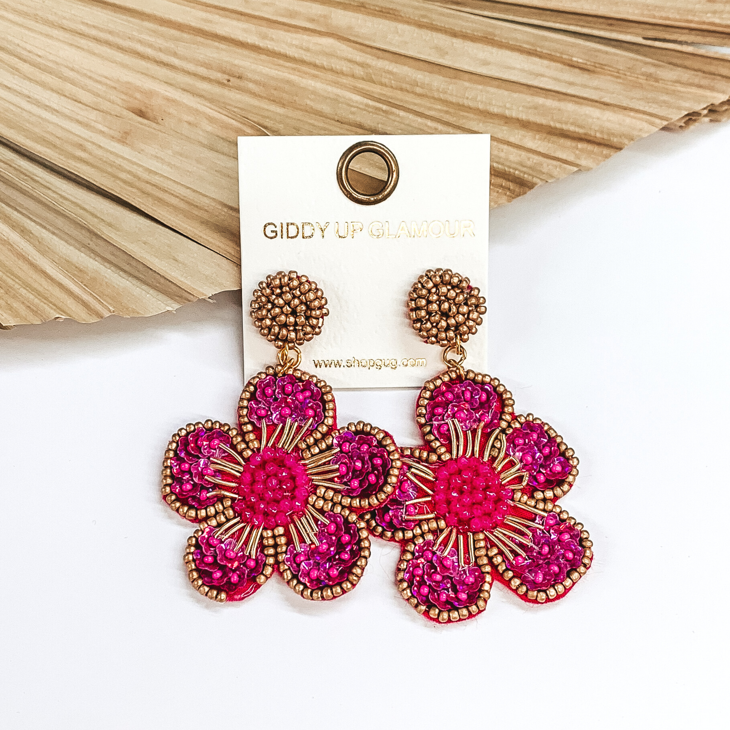 Gold beaded circle post back earrings. There is a hanging beaded flower pendant from the gold stud earrings. The flower pendant is fuchsia with a gold outline and detailing. These earrings are pictured on a white background with a dried palm leaf in the background. 