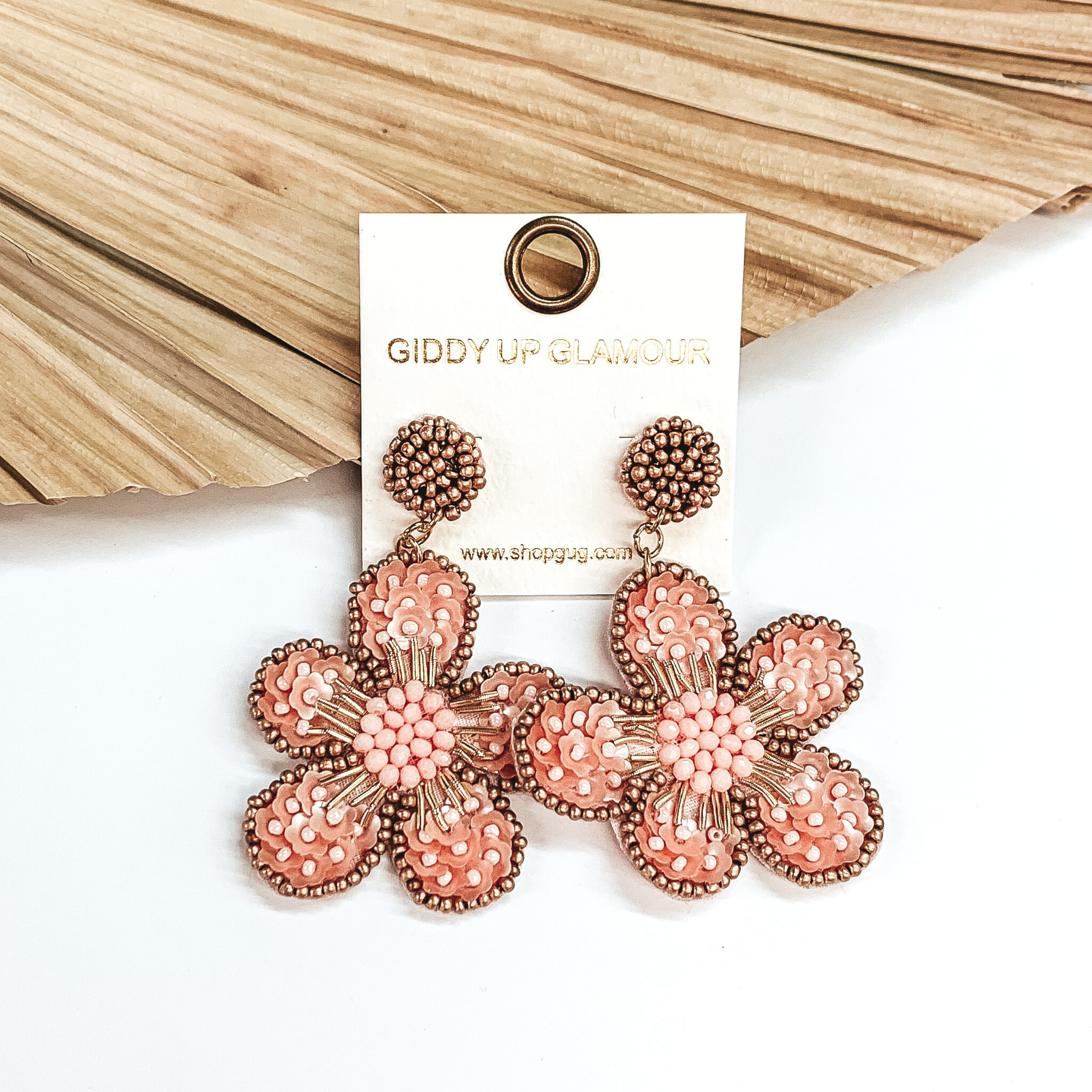 Gold beaded circle post back earrings. There is a hanging beaded flower pendant from the gold stud earrings. The flower pendant is baby pink with a gold outline and detailing. These earrings are pictured on a white background with a dried palm leaf in the background. 