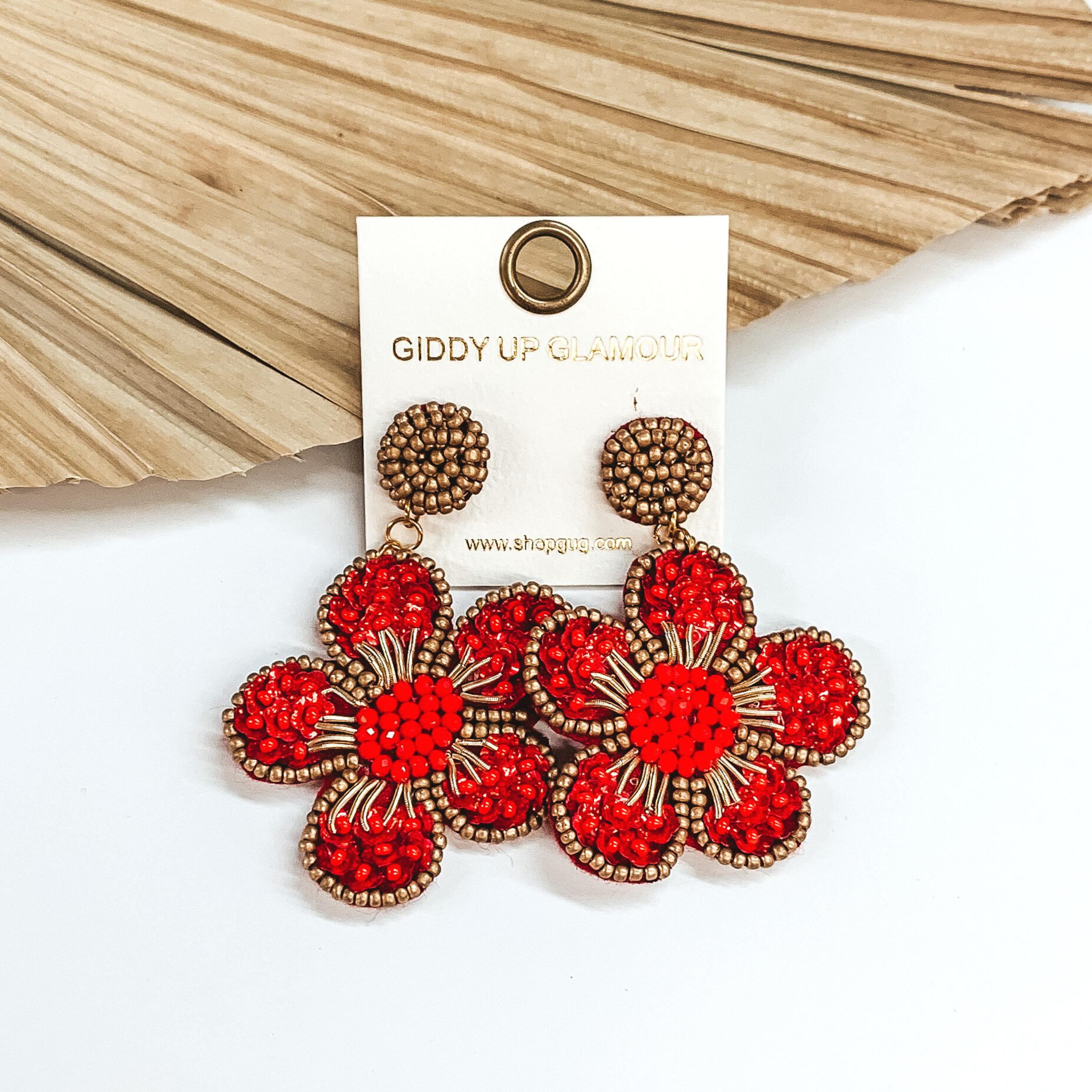 Gold beaded circle post back earrings. There is a hanging beaded flower pendant from the gold stud earrings. The flower pendant is red with a gold outline and detailing. These earrings are pictured on a white background with a dried palm leaf in the background. 