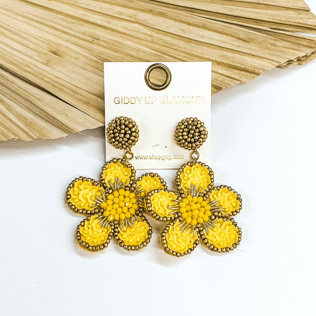 Gold beaded circle post back earrings. There is a hanging beaded flower pendant from the gold stud earrings. The flower pendant is yellow with a gold outline and detailing. These earrings are pictured on a white background with a dried palm leaf in the background. 
