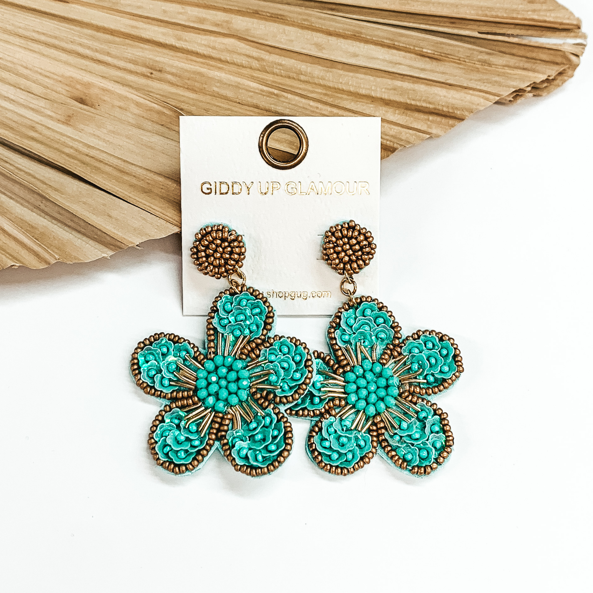 Gold beaded circle post back earrings. There is a hanging beaded flower pendant from the gold stud earrings. The flower pendant is turquoise with a gold outline and detailing. These earrings are pictured on a white background with a dried palm leaf in the background. 