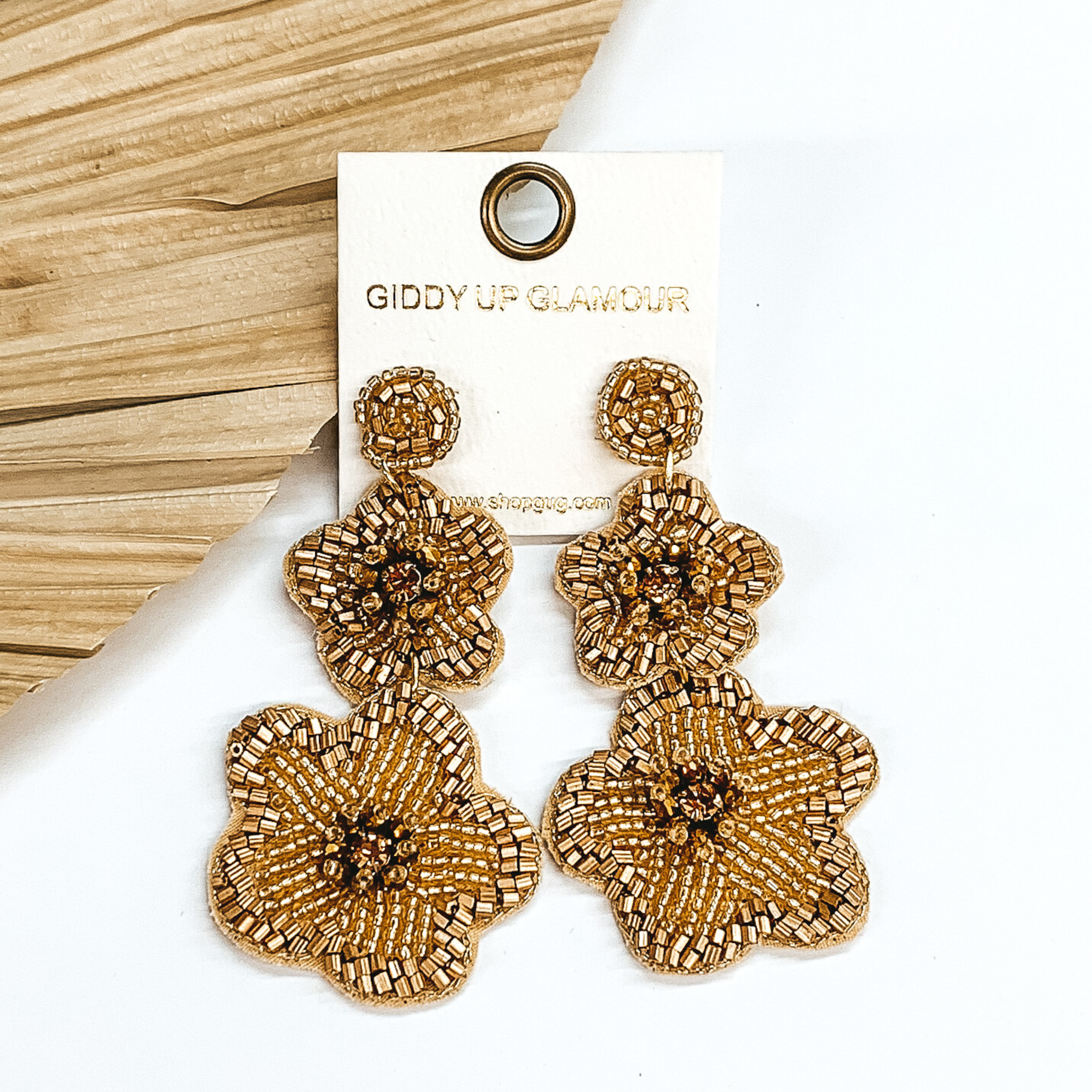 Gold beaded circle post back earrings. There is a two hanging beaded flower pendant from the gold stud earrings. The flower pendants are gold with a gold outline and detailing. These earrings are pictured on a white background with a dried palm leaf in the background. 