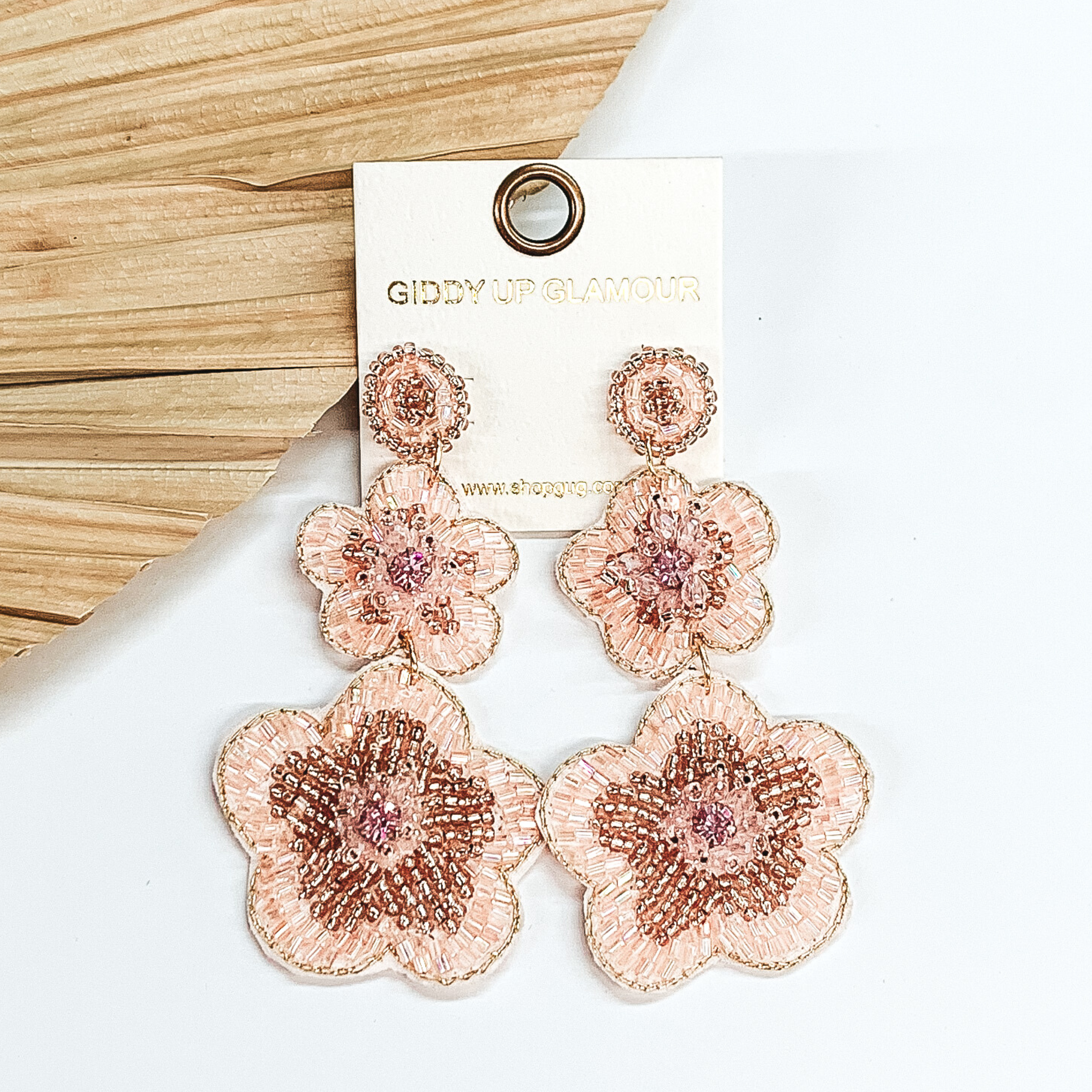 Gold beaded circle post back earrings. There is a two hanging beaded flower pendant from the gold stud earrings. The flower pendants are light pink with a rose gold outline and detailing. These earrings are pictured on a white background with a dried palm leaf in the background. 