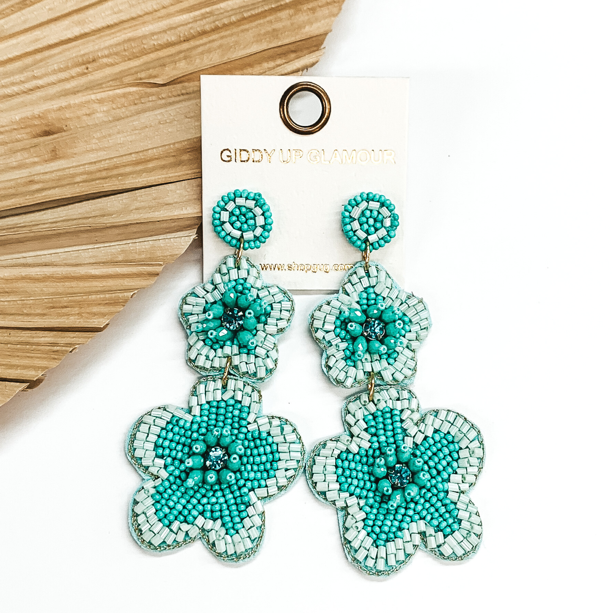 Gold beaded circle post back earrings. There is a two hanging beaded flower pendant from the gold stud earrings. The flower pendants are turquoise with a light aqua outline and detailing. These earrings are pictured on a white background with a dried palm leaf in the background. 