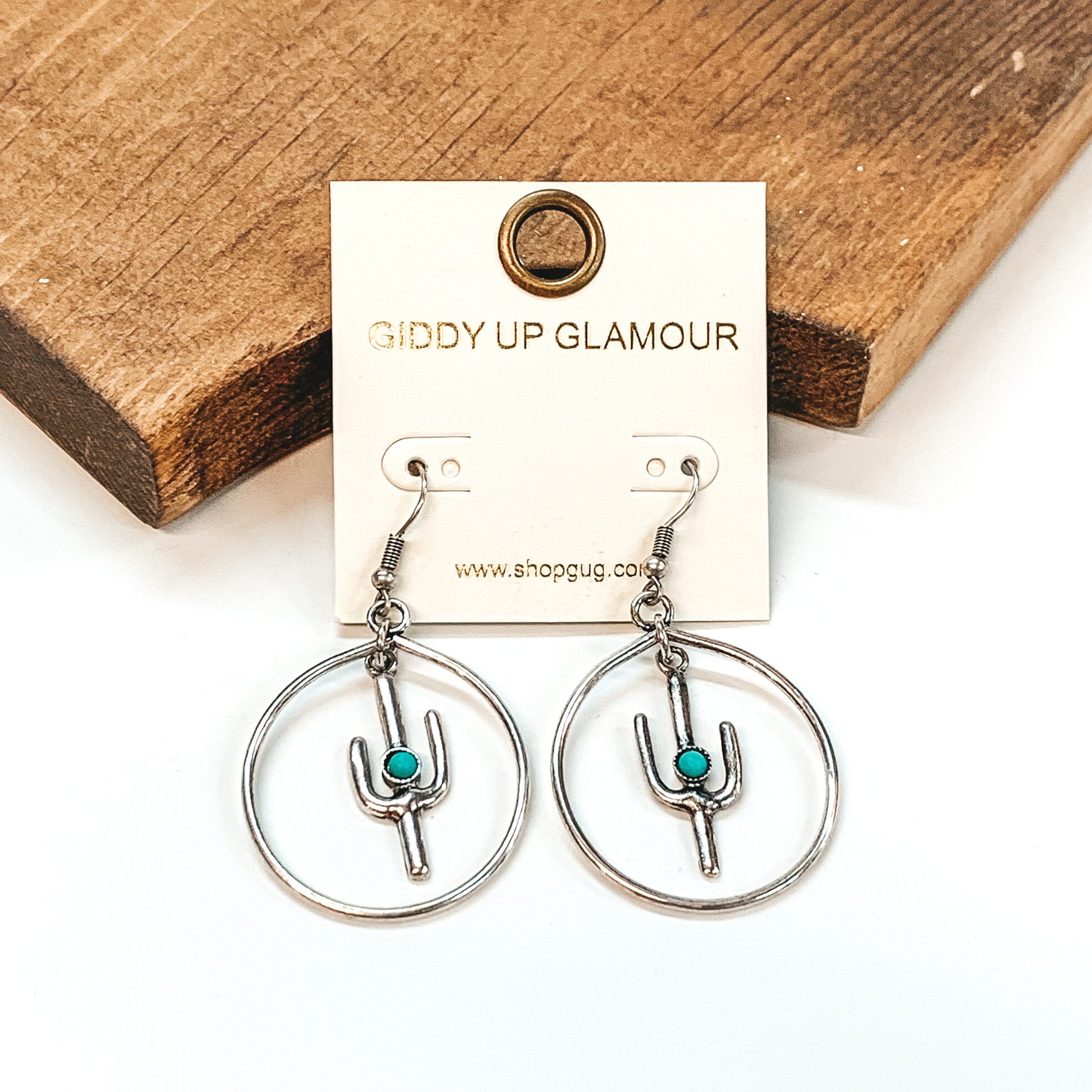 Silver fish hook earrings with a circle drop pendant. Hanging in the middle of the circle is a cactus pendant that has a tiny turquoise stone. These earrings are pictured on a white background in front of a brown block.  