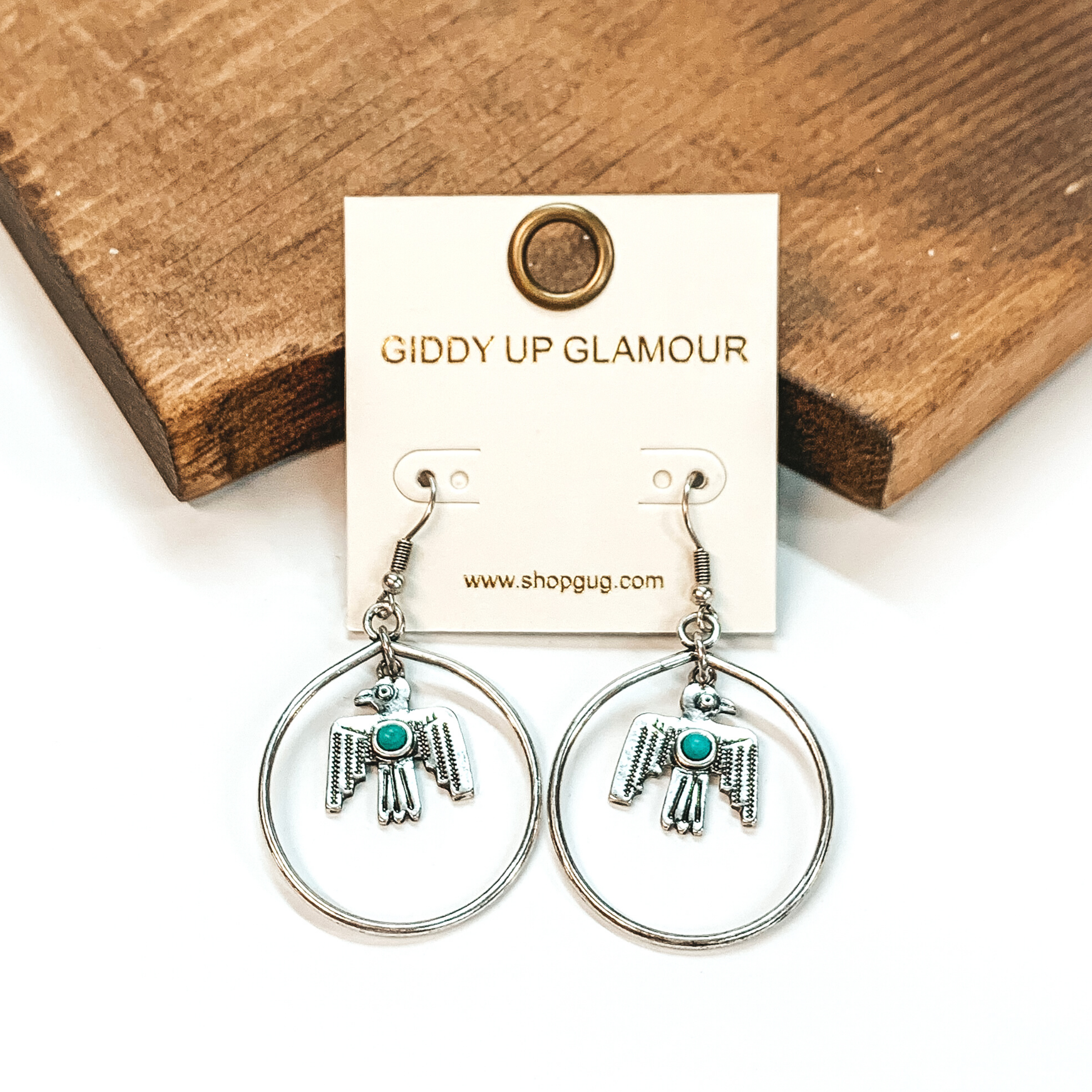 Silver fish hook earrings with a circle drop pendant. Hanging in the middle of the circle is a thunderbird pendant that has a tiny turquoise stone. These earrings are pictured on a white background in front of a brown block. 