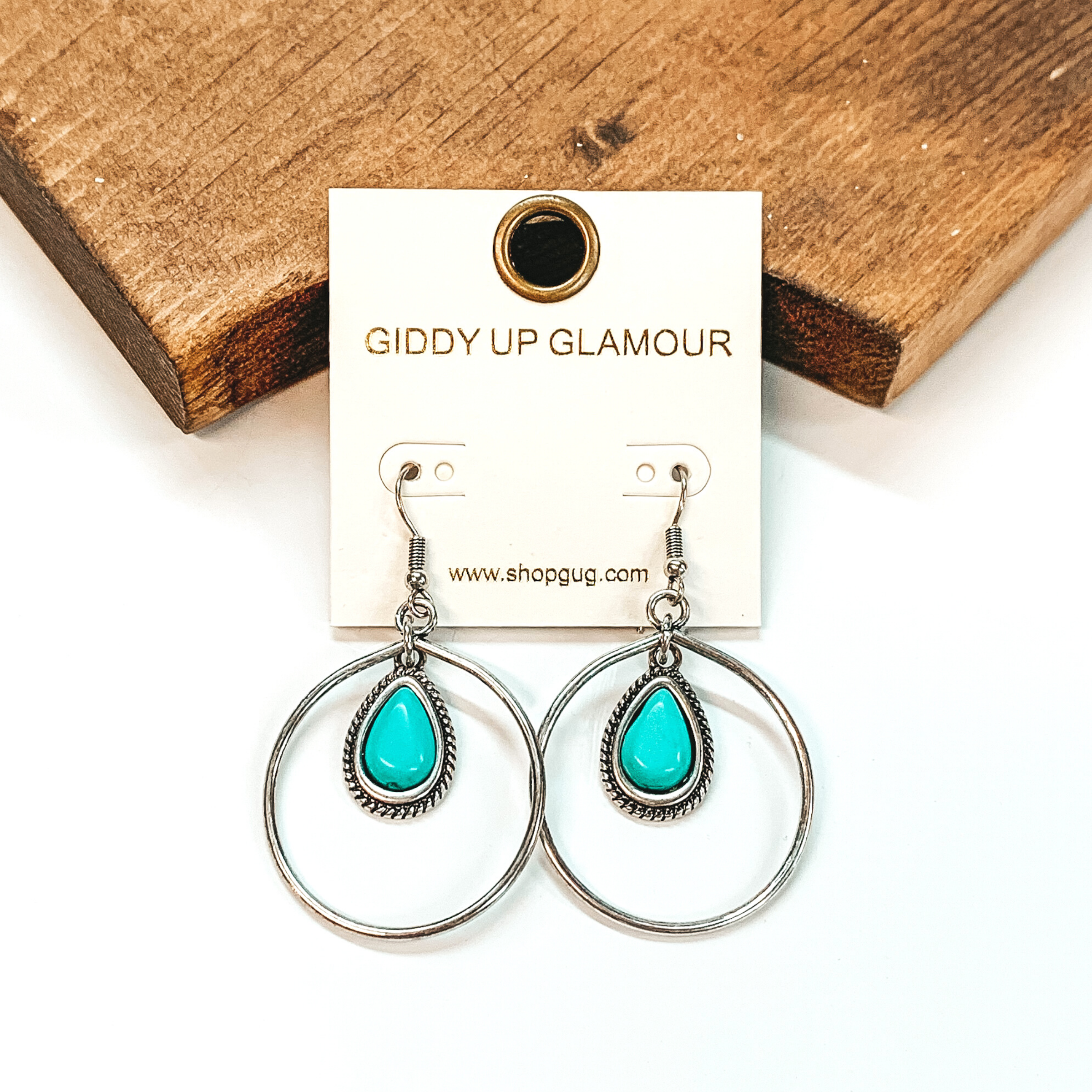 Silver fish hook earrings with a circle drop pendant. Hanging in the middle of the circle is a teardrop turquoise stone pendant. These earrings are pictured on a white background in front of a brown block.  
