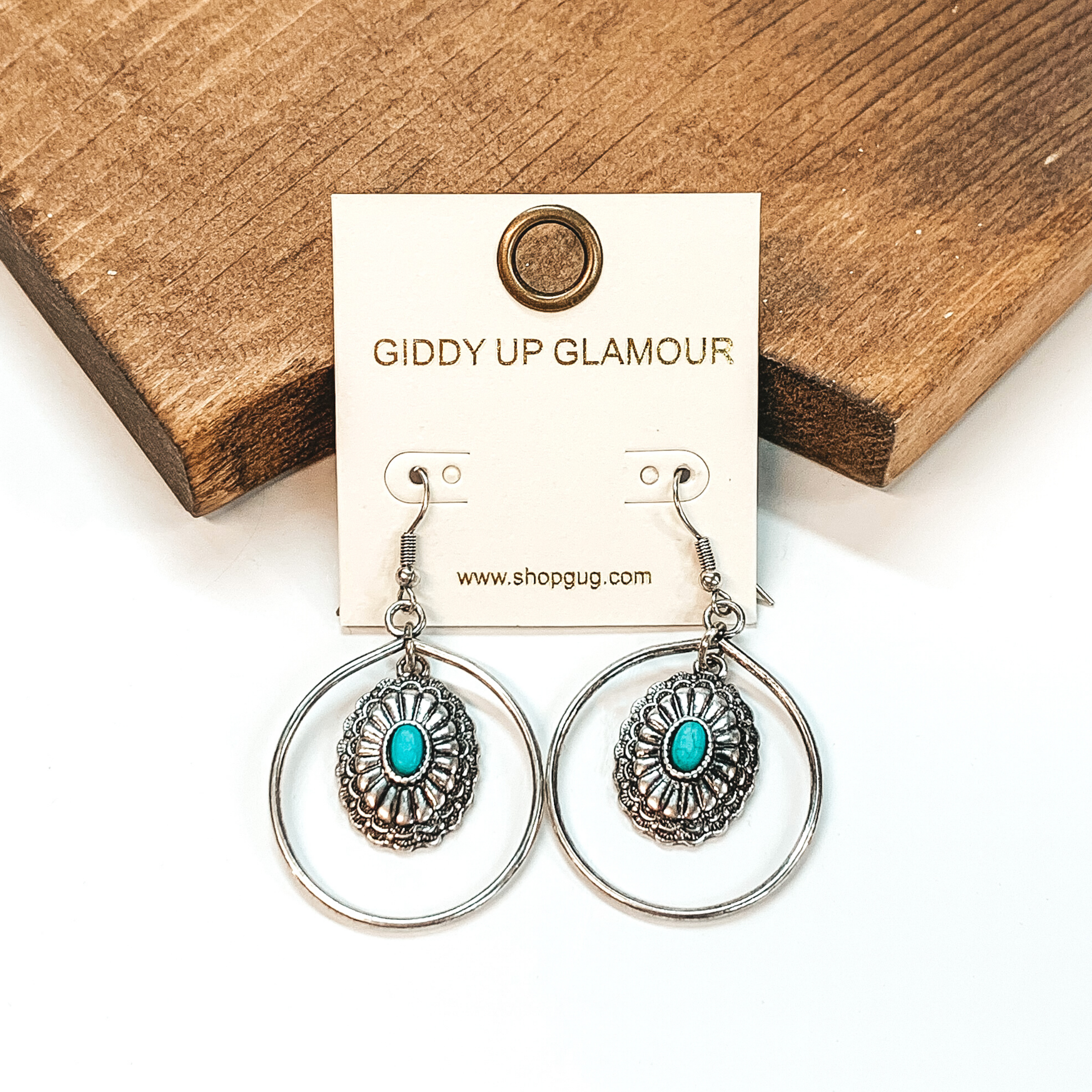  Silver fish hook earrings with a circle drop pendant. Hanging in the middle of the circle is a concho pendant that has a tiny turquoise center stone. These earrings are pictured on a white background in front of a brown block.