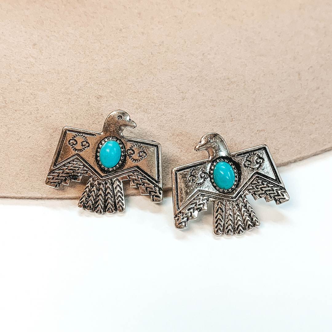 Silver thunderbird stud earrings with a center, oval, turquoise stone. These earrings are pictured on a white and beige background.