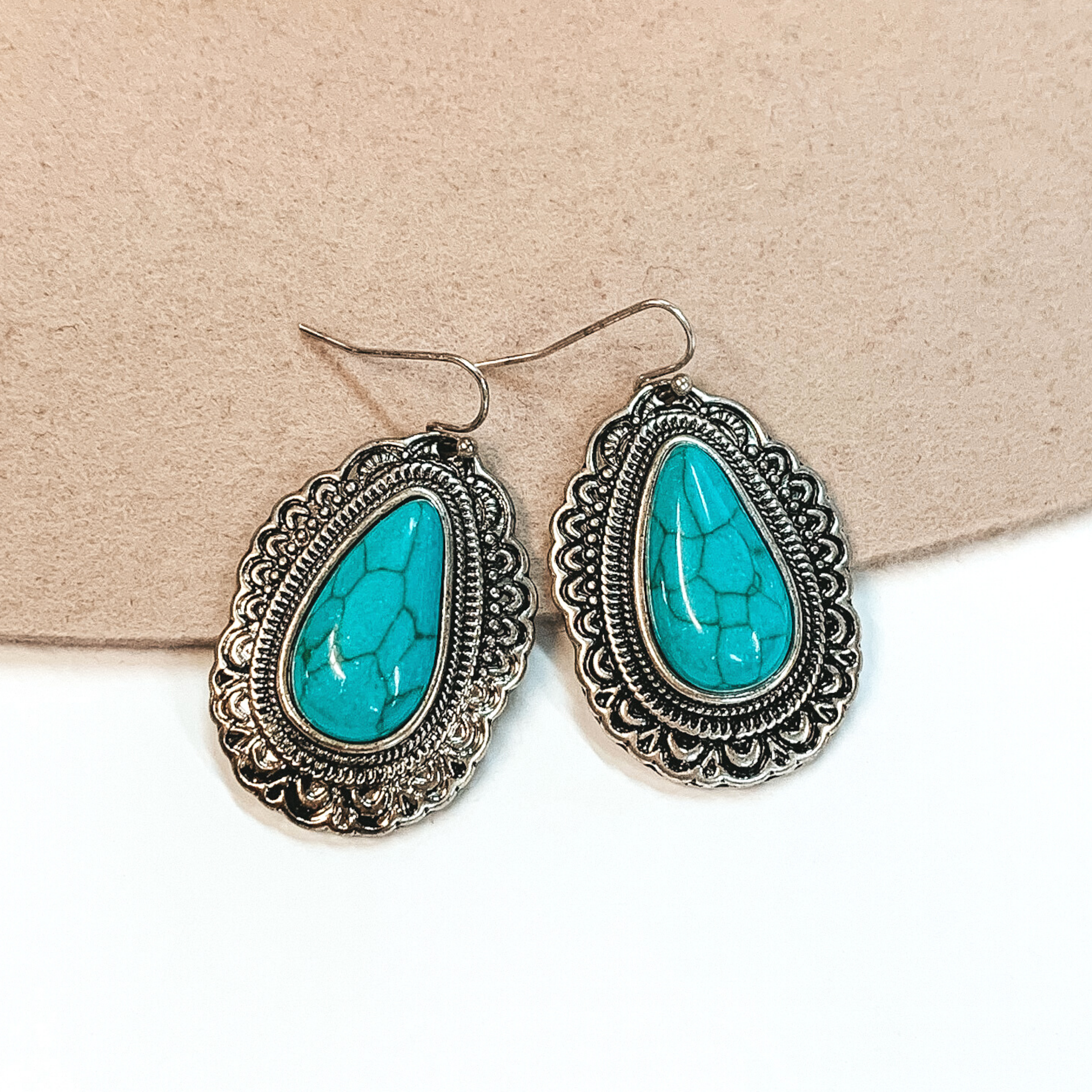 These are silver teardrop earrings in silver fish hooks with a teardrop turquoise stone. There are engraved detailing around the stone. These earrings are pictured on a white and beige background. 