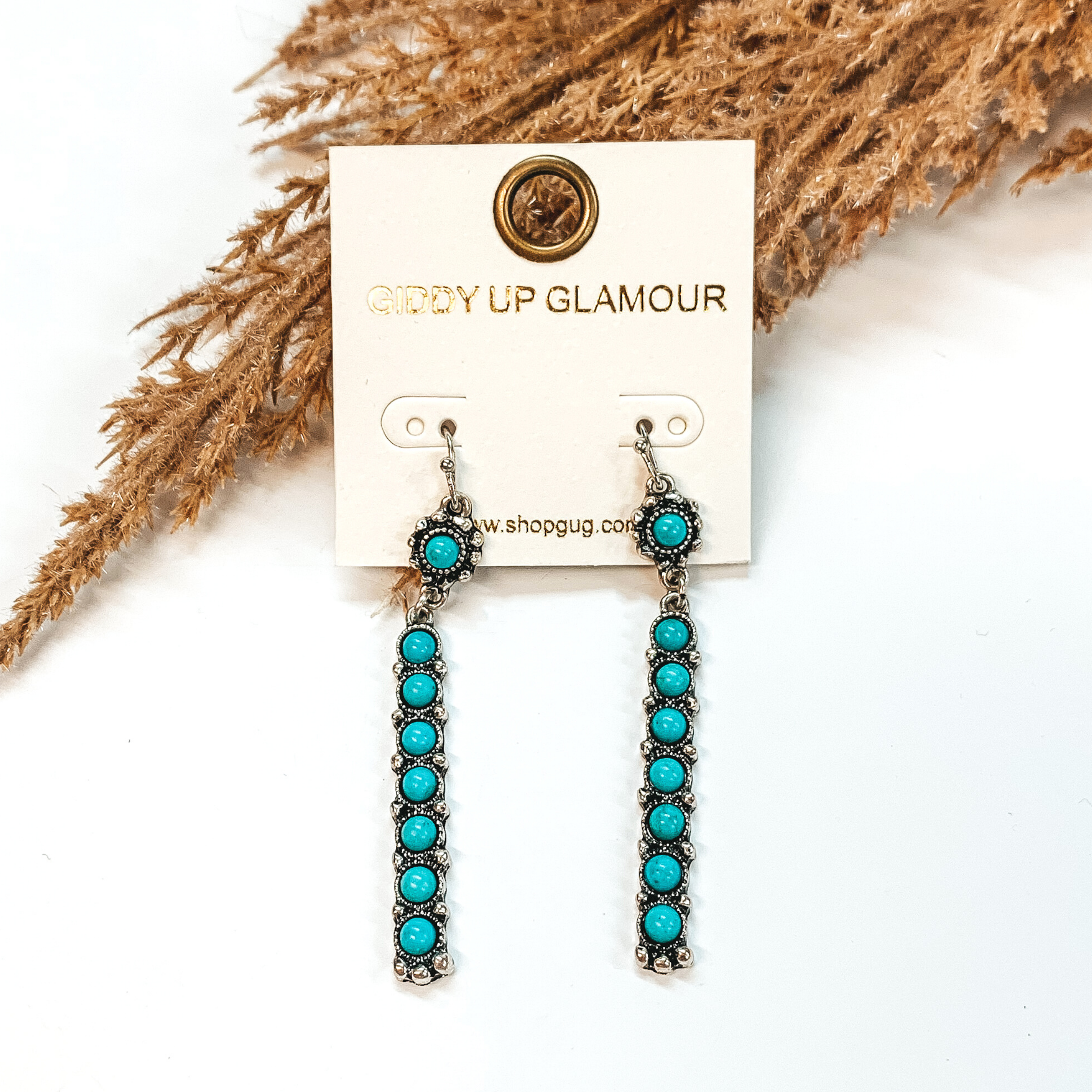On silver fish hook earrings there is a small circle drop pendant with a turquoise stone. Hanging from the bottom is a rectangle bar that has an inlay of circle turquoise stones. These earrings are pictured on a white background in front of brown floral decor.