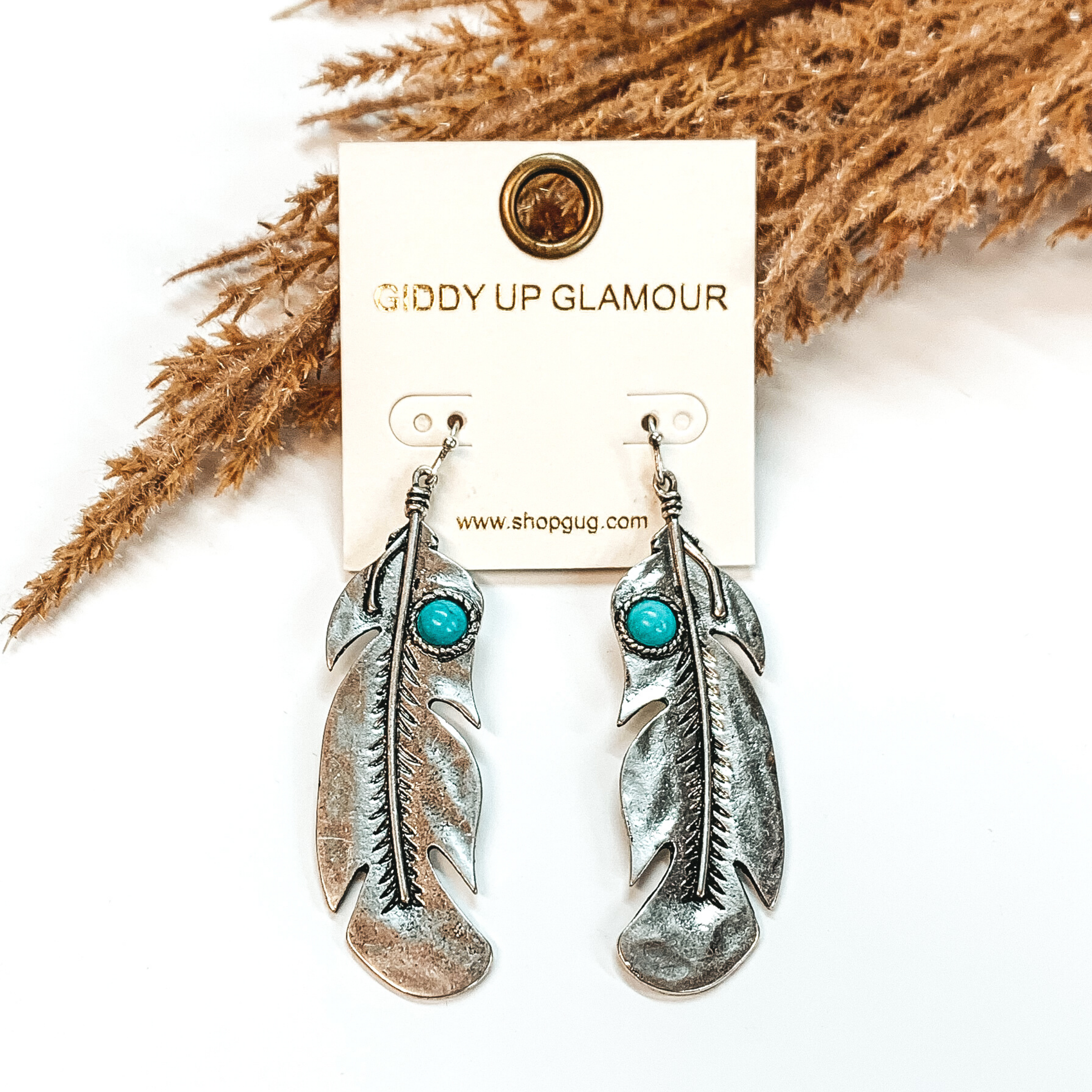 On silver fish hook earrings is a silver feather pendant with engraved deather detaileing. Towards the top of each feather, there is a small, circle turquoise stone. These earrings are pictured on a white background in front of brown floral decor.