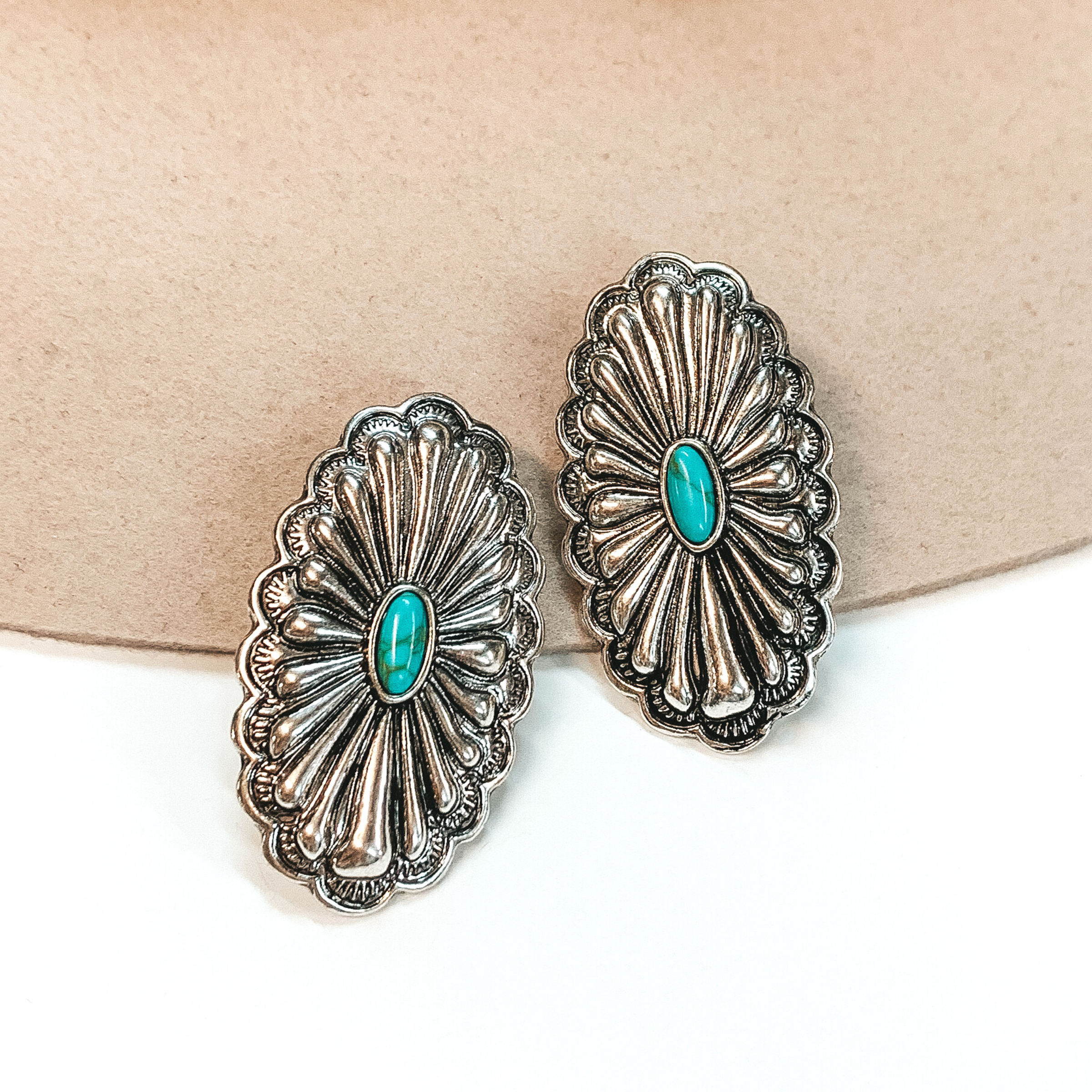 Silver, oval concho earrings with a western design. There is a center, oval turquoise stone. These earrings are pictured on a white and beige background. 