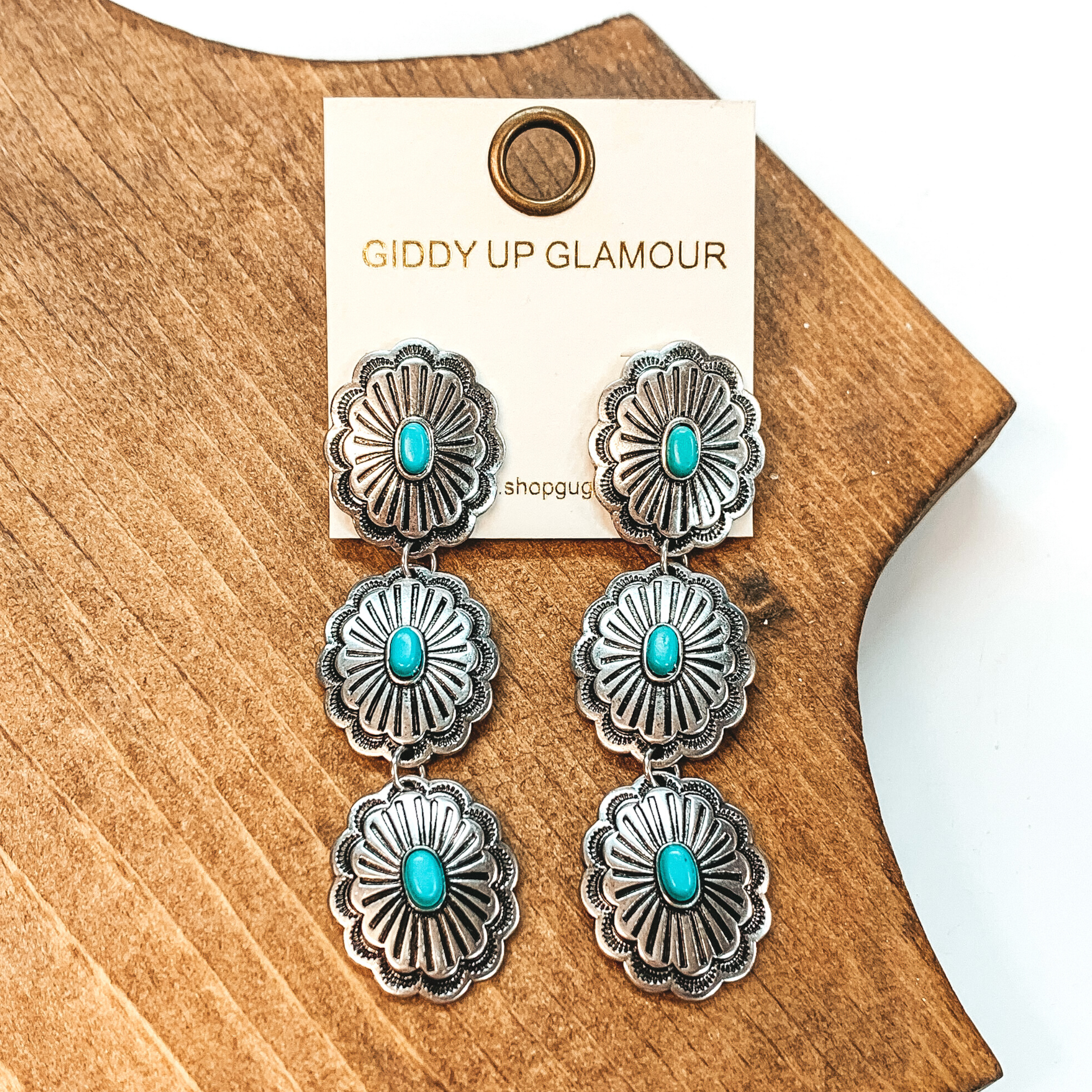 Silver, oval concho dangle earrings with a western design. At the center of all three conchos is a center, oval turquoise stone. These earrings are pictured on a white background in front of a brown block. 