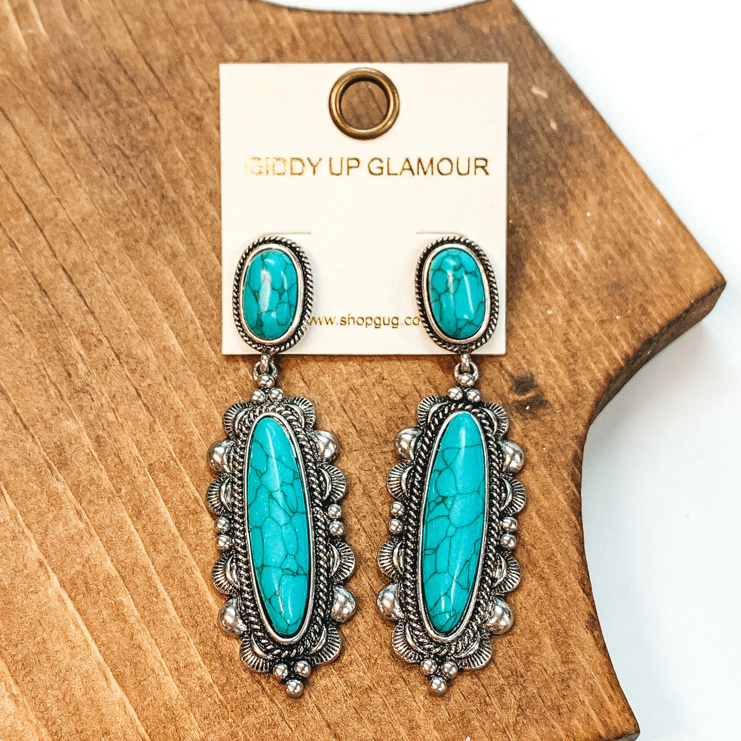 Turquoise, oval post back earrings with a silver outline. At the bottom of the earrings is a oval pendant. The center has an oval, turquoise stone with aa western, silver outline. These earrings are pictured laying on a brown block on a white background.