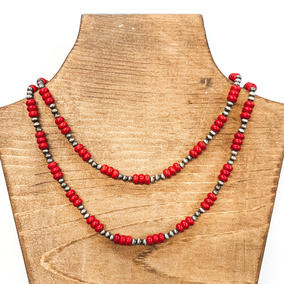 Two strand necklace that includes silver bead segments with bigger, red bead segments. This necklace is pictured on a brown necklace holder on a white background.