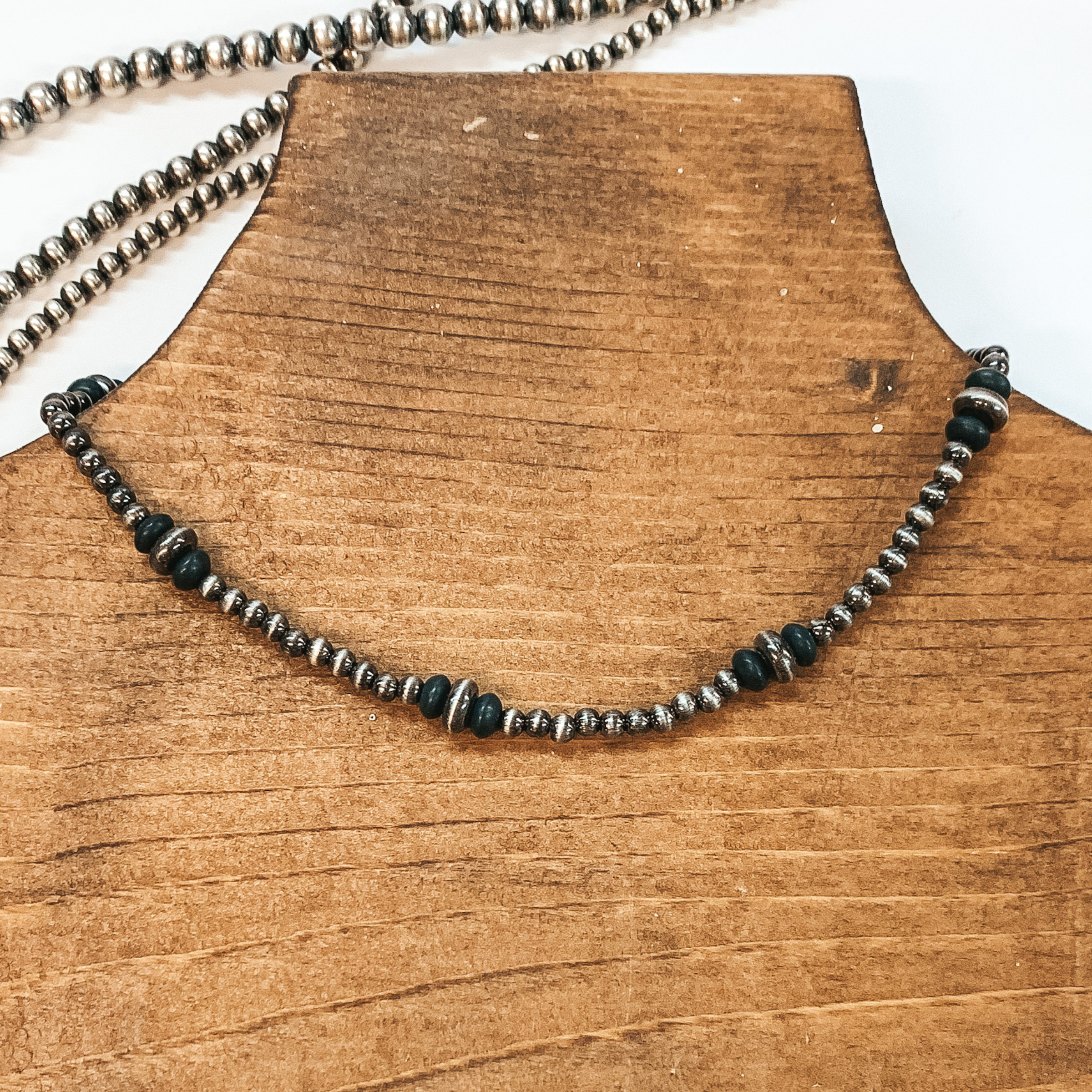 This is a silver beaded necklace with spacers of two different size saucer beads in black. This necklace is pictured on a brown necklace holder on a white background with silver beads at the top.