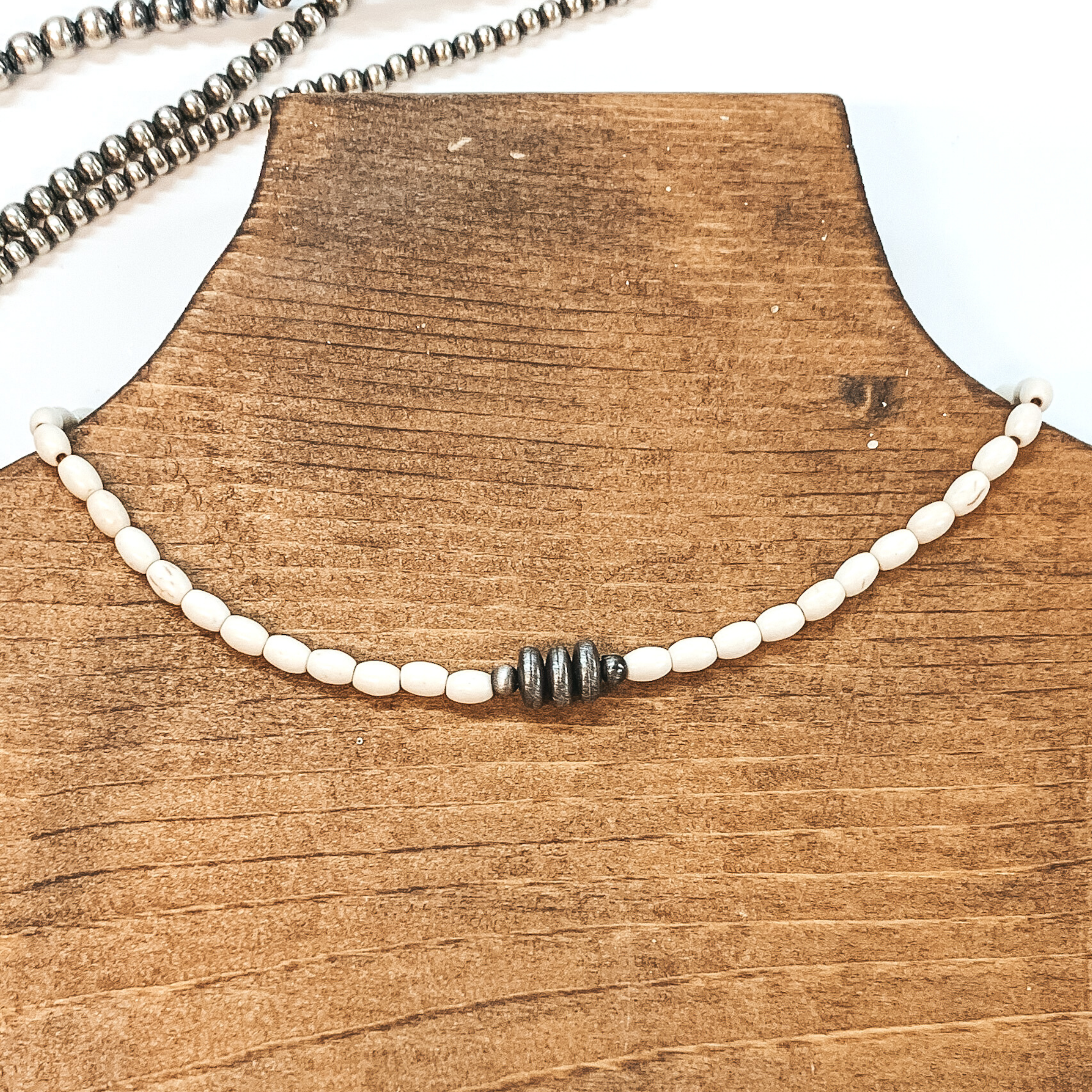Ivory beaded necklace with silver, saucer beads in the center. This necklace is pictured on a brown necklace holder on a white background with silver beads in the background. 