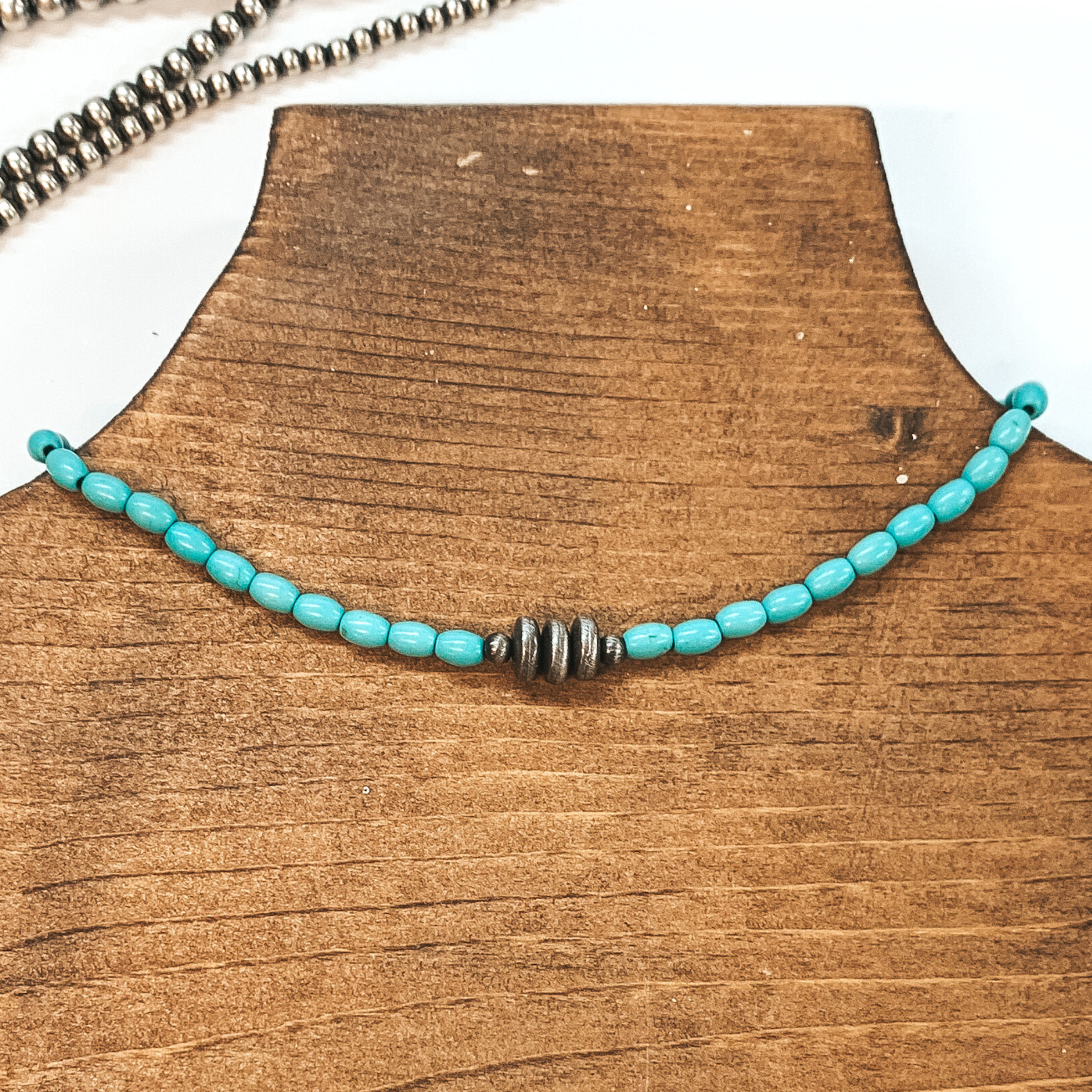 Turquoise beaded necklace with silver, saucer beads in the center. This necklace is pictured on a brown necklace holder on a white background with silver beads in the background.  