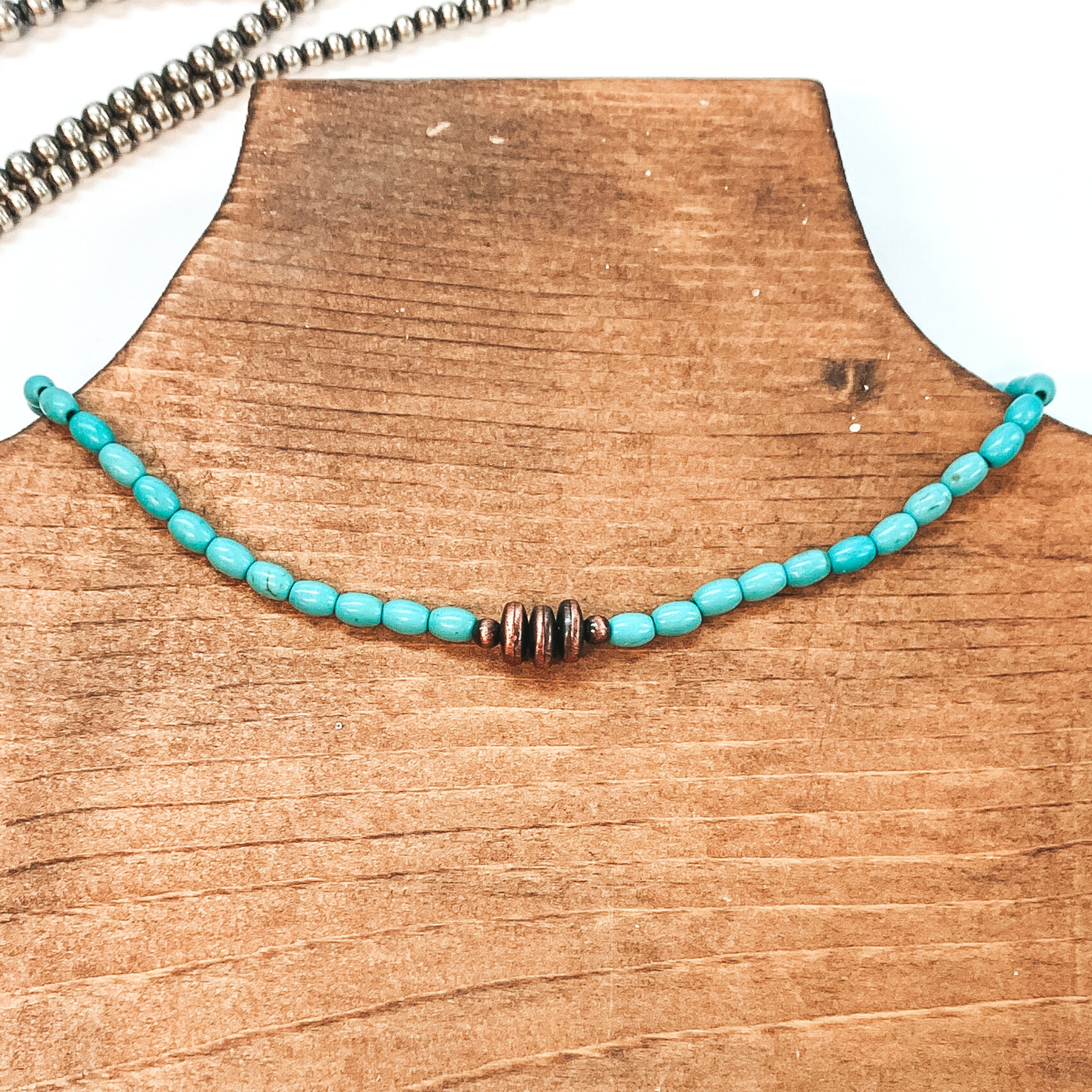 Turquoise beaded necklace with copper, saucer beads in the center. This necklace is pictured on a brown necklace holder on a white background with silver beads in the background. 