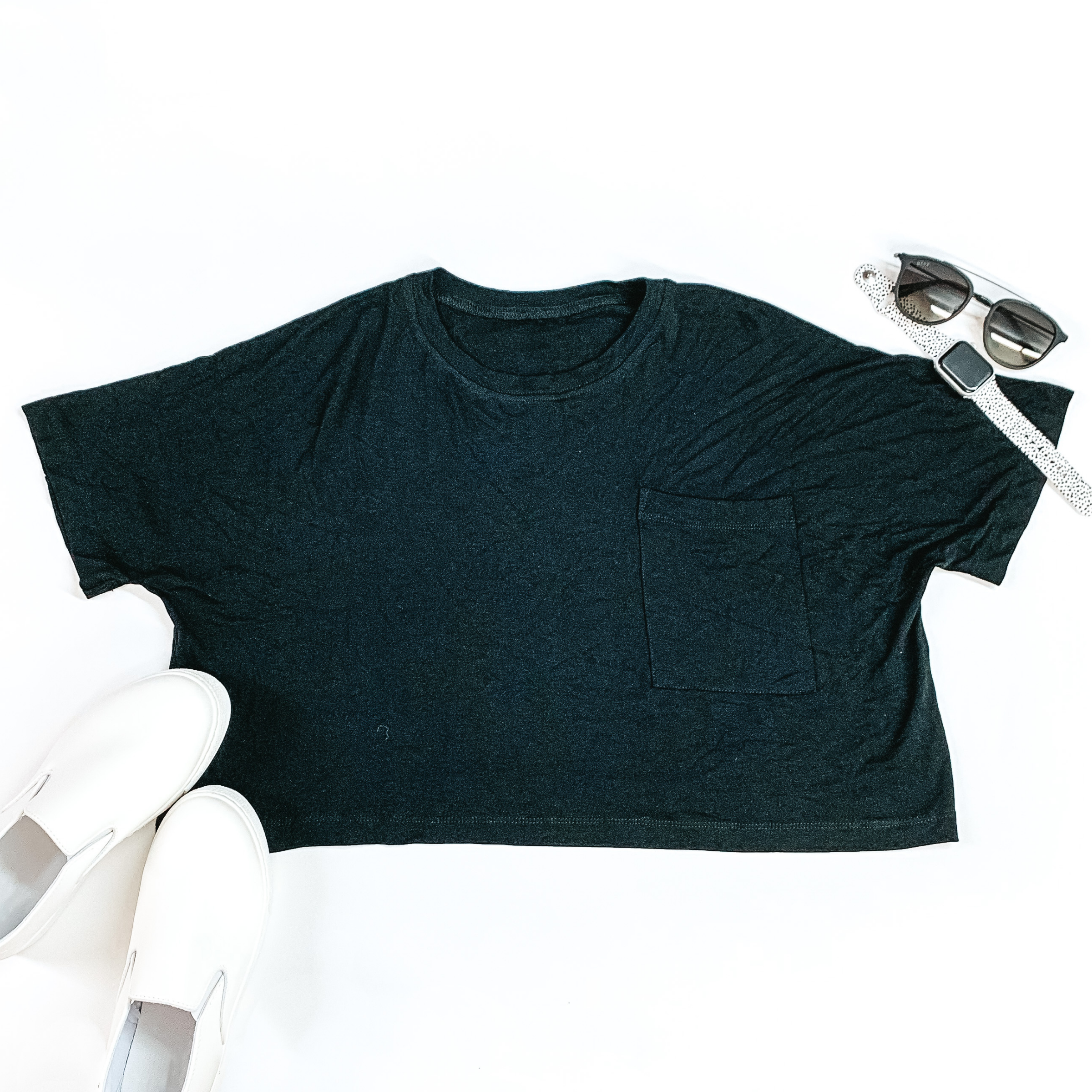 Black cropped short sleeve tee with a front pocket and a scoop neck. This tee is pictured on a white background with with slip on and sunglasses in the picture. 